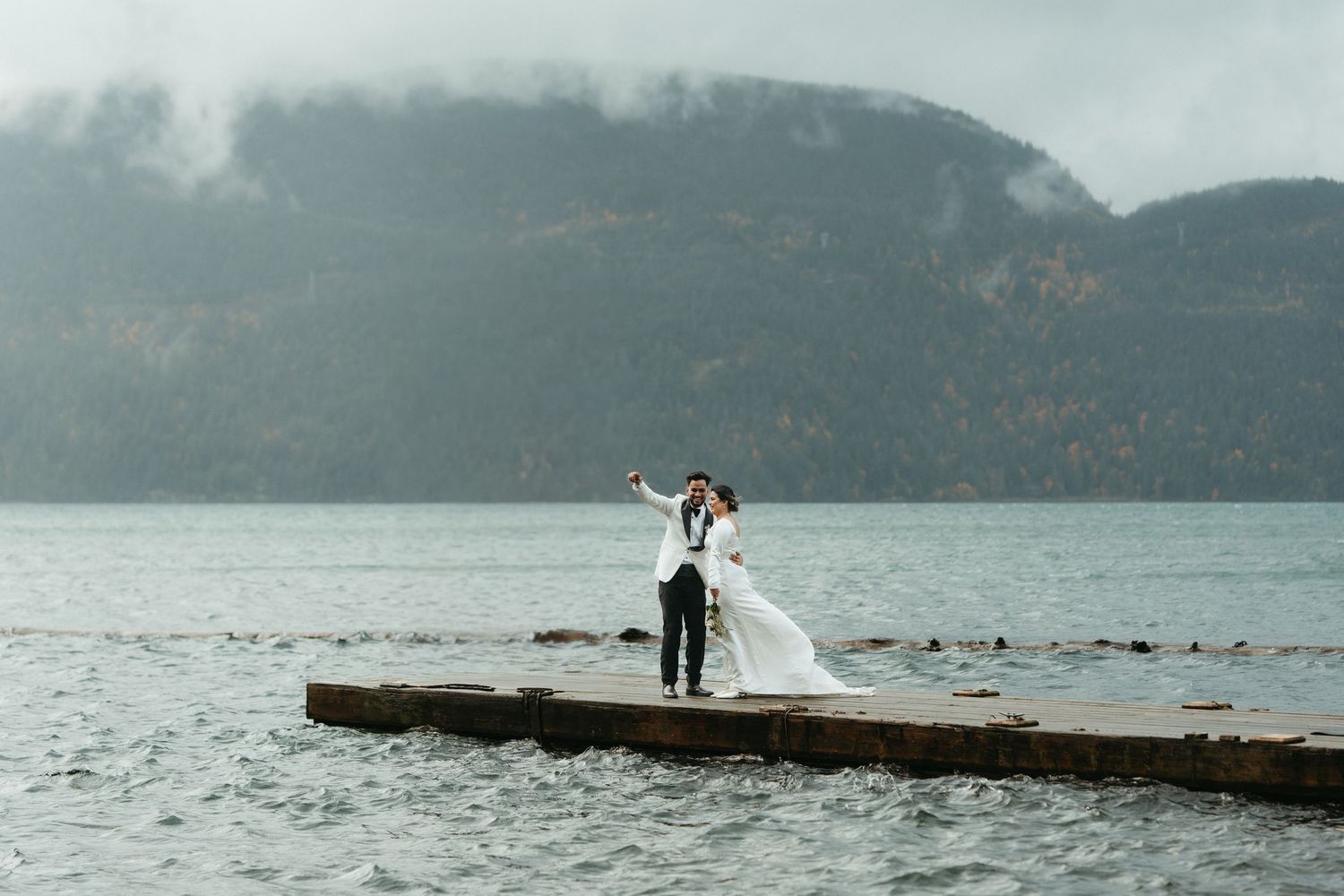 A couple stands on a rocky ocean pier with mountains in the background on an overcast day.