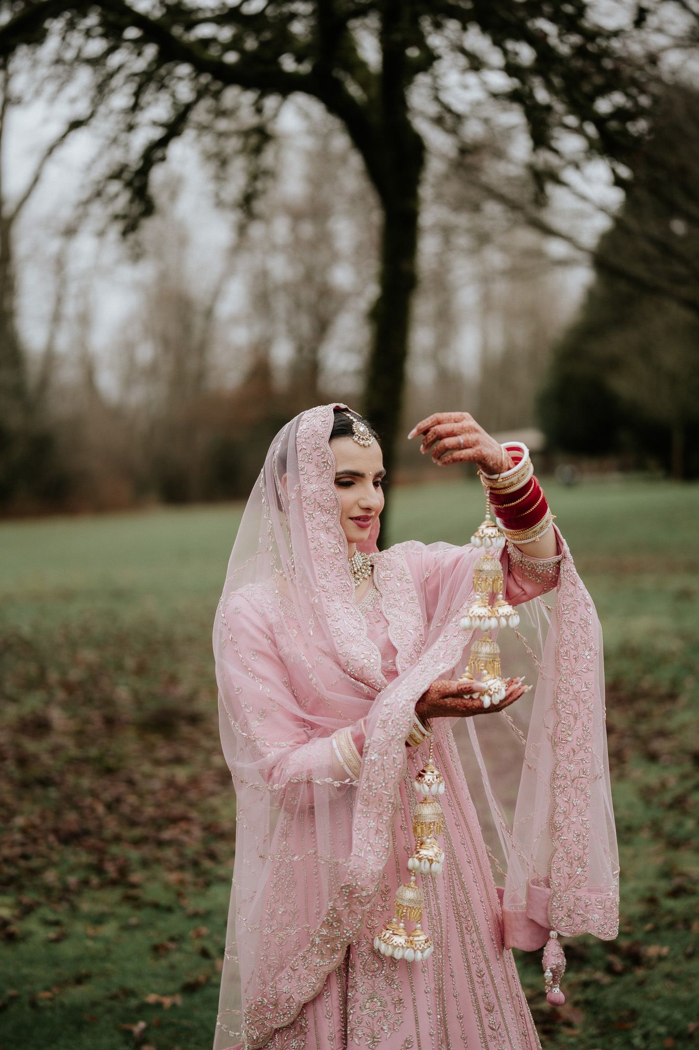 Traditional bride in pink embroidered dress and veil poses gracefully with jewelry in an outdoor garden setting.