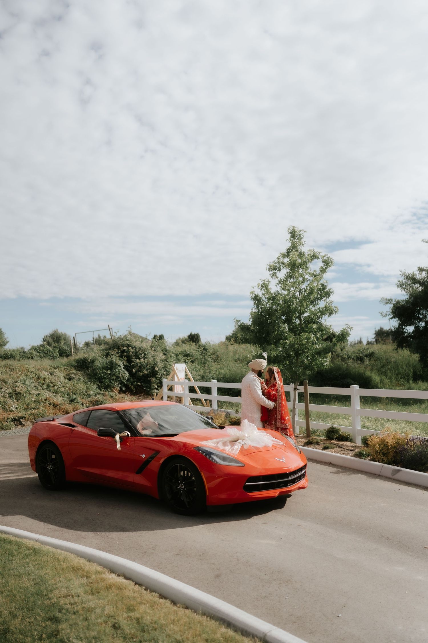 Couple in traditional wedding attire posing behind a red Corvette on a scenic road with white fence and greenery.
