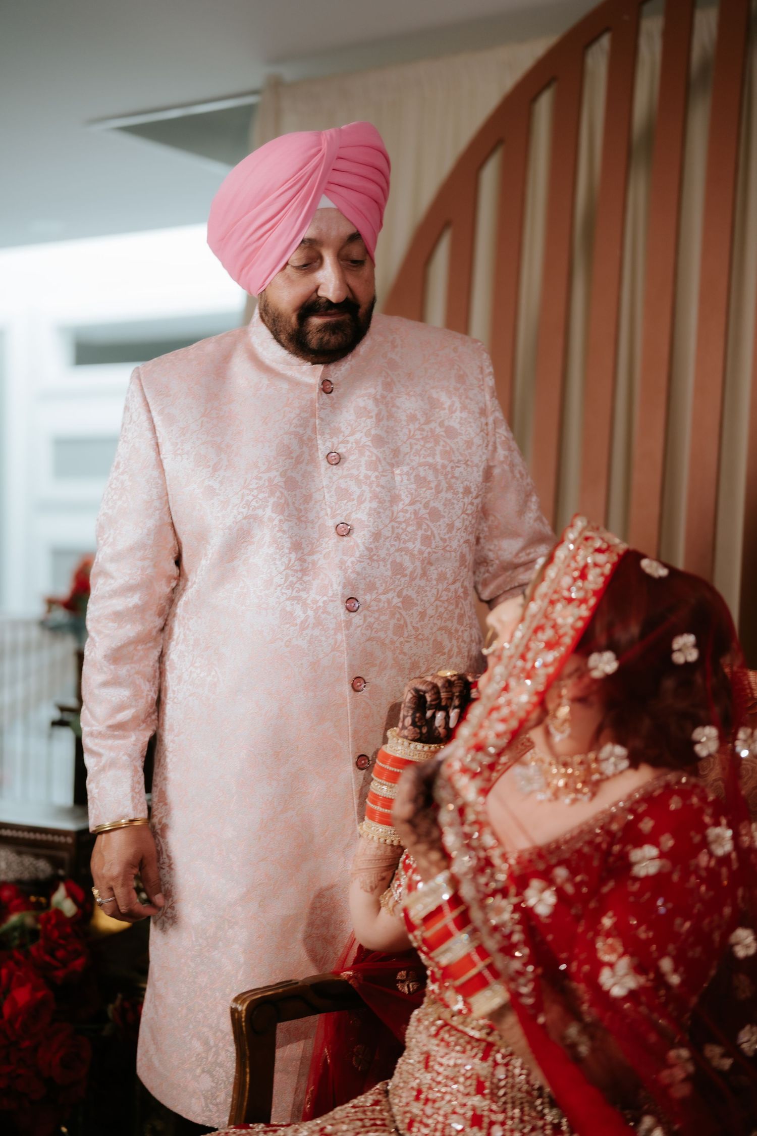 Sikh groom in pink turban and sherwani stands beside bride in red lehenga during traditional Indian wedding ceremony.