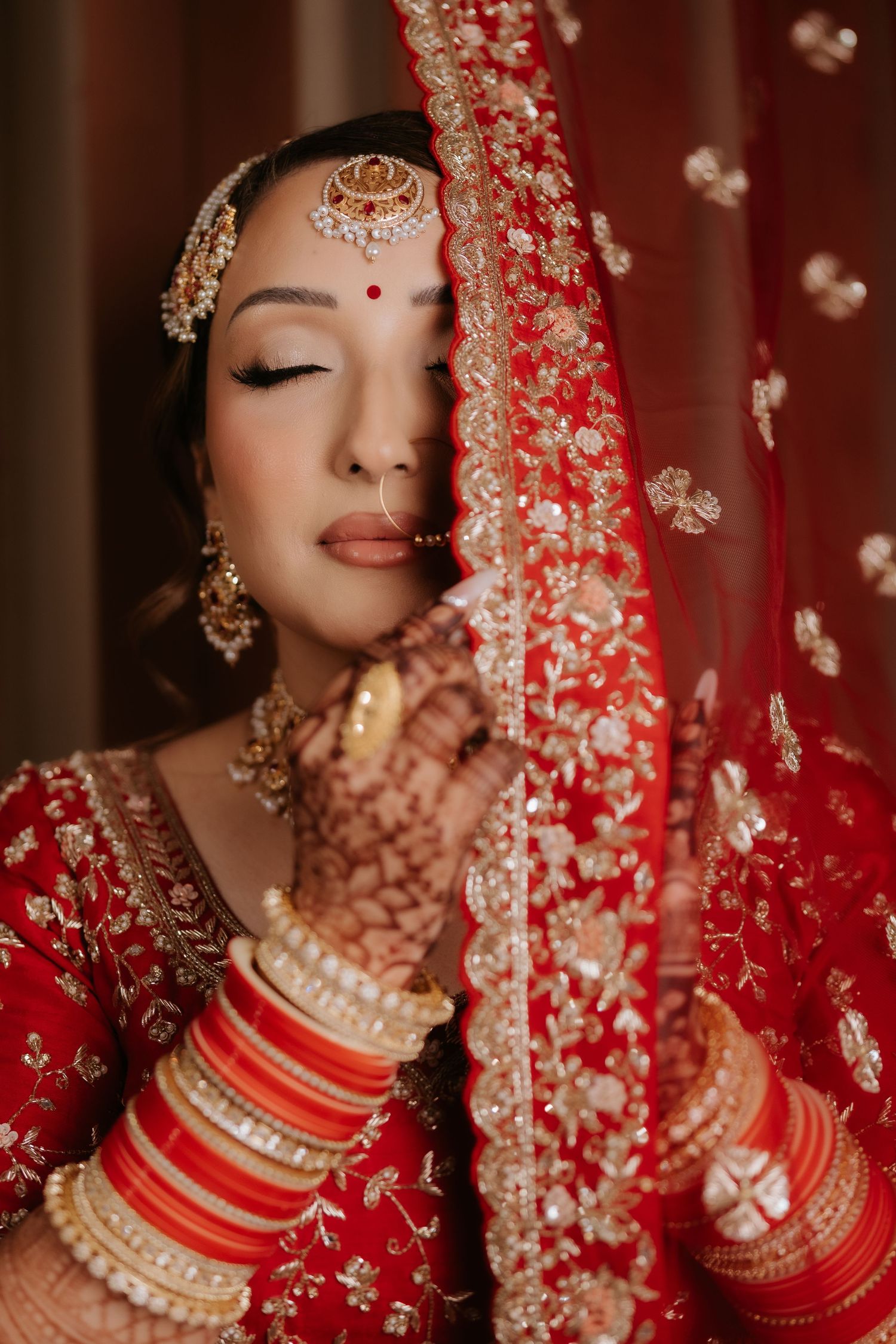 Indian bride in red lehenga with gold embroidery, wearing traditional jewelry and bangles, holding red dupatta near her face.