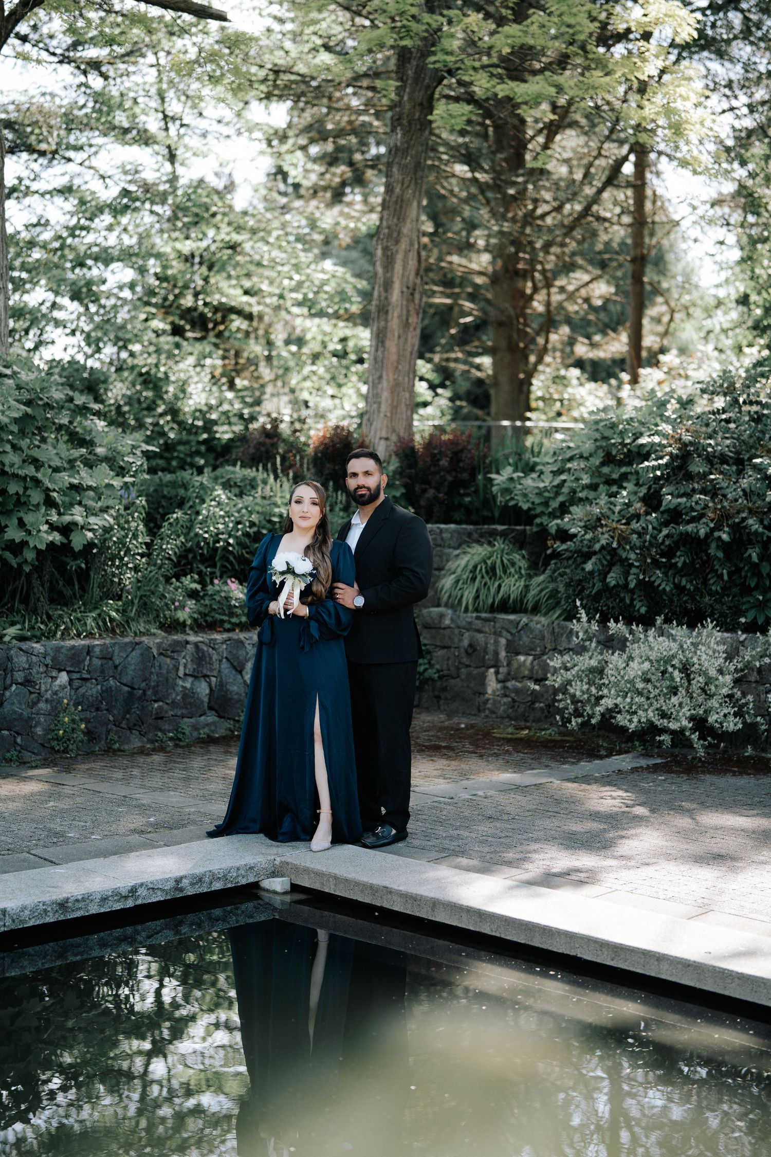 Elegant couple in navy formal wear posing by a reflective garden pond surrounded by lush greenery.