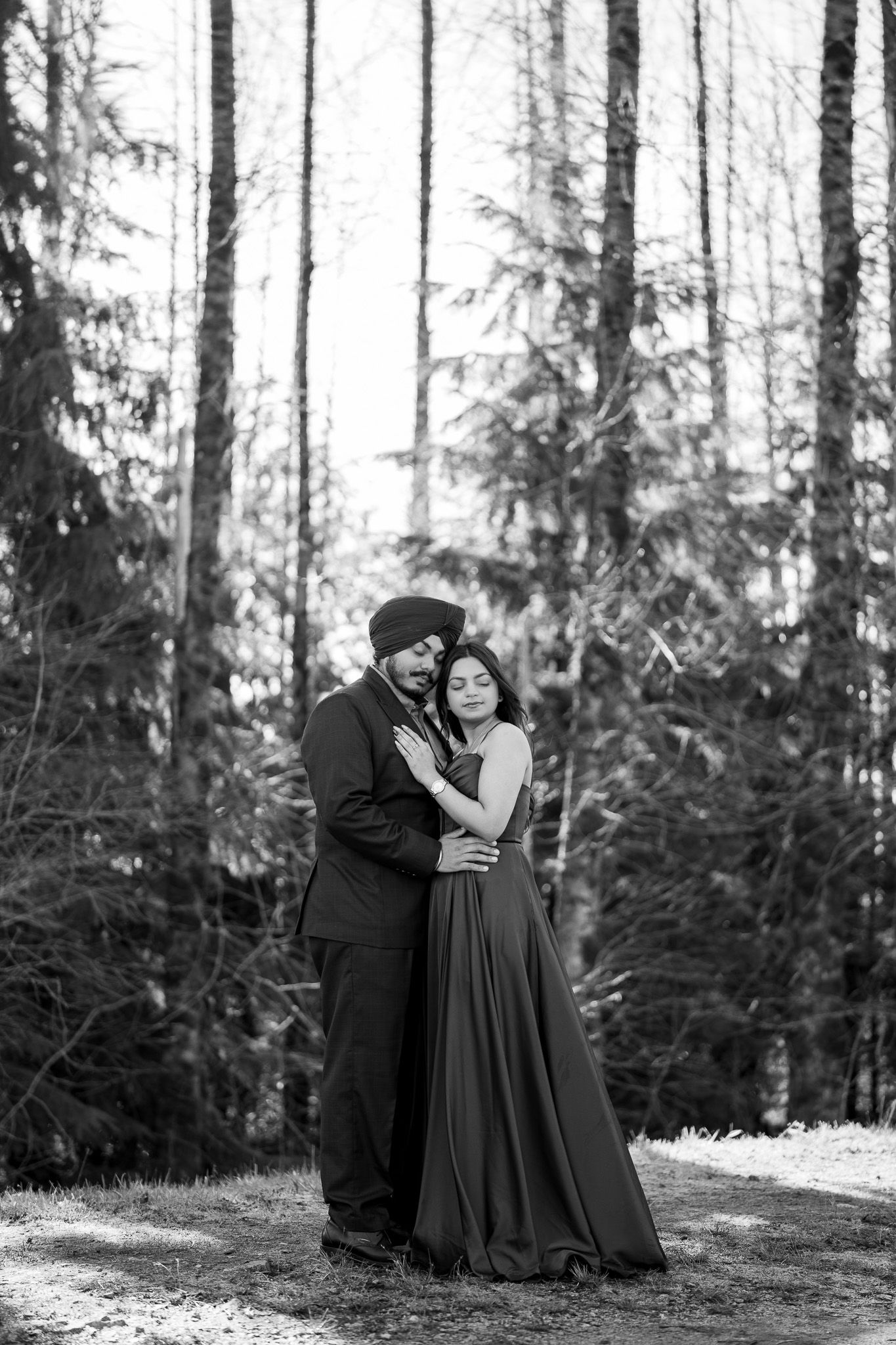 A romantic couple in formal attire embraces in a forest setting with evergreen trees in the background.