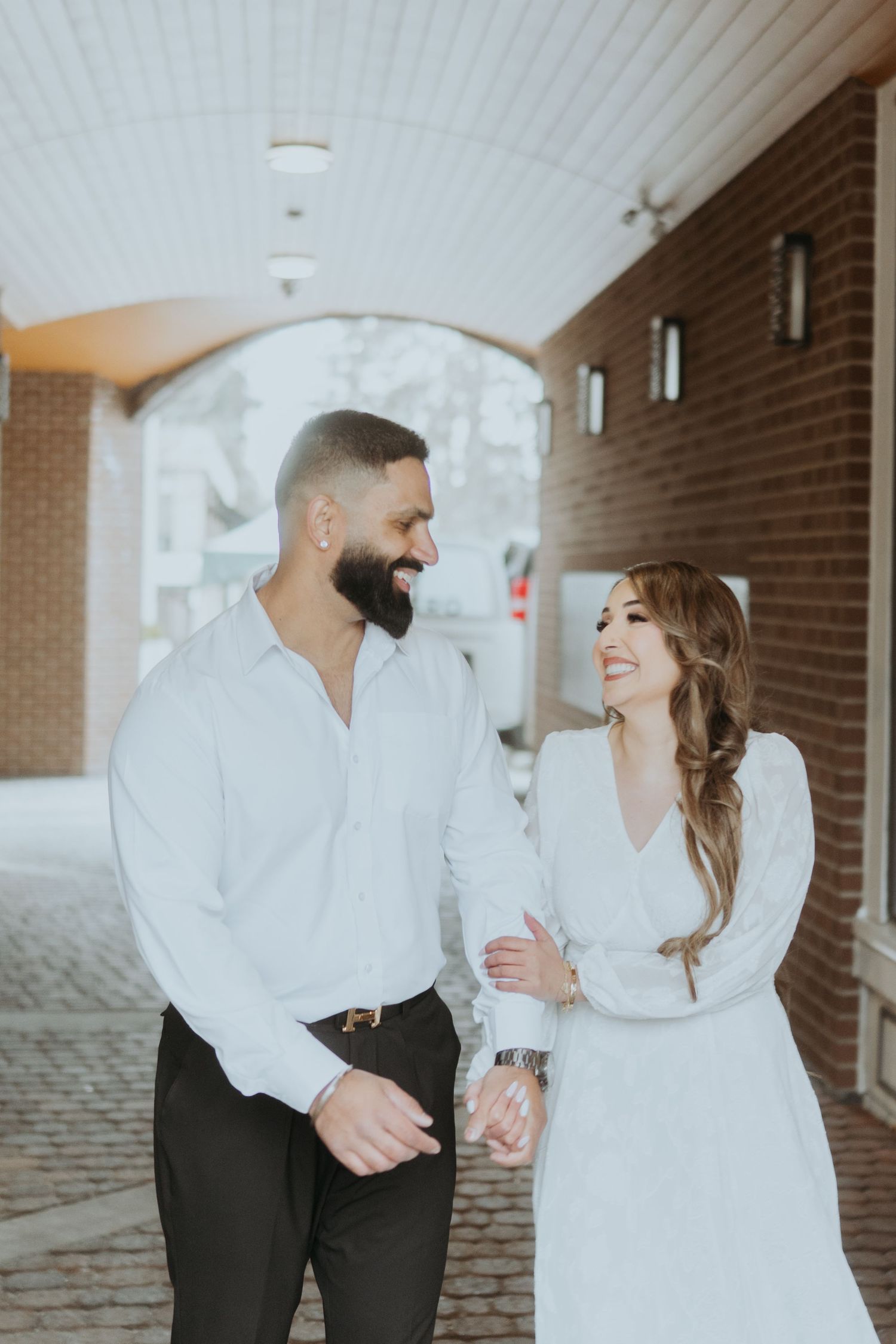 A couple in white and black attire shares a tender moment in a brick hallway.