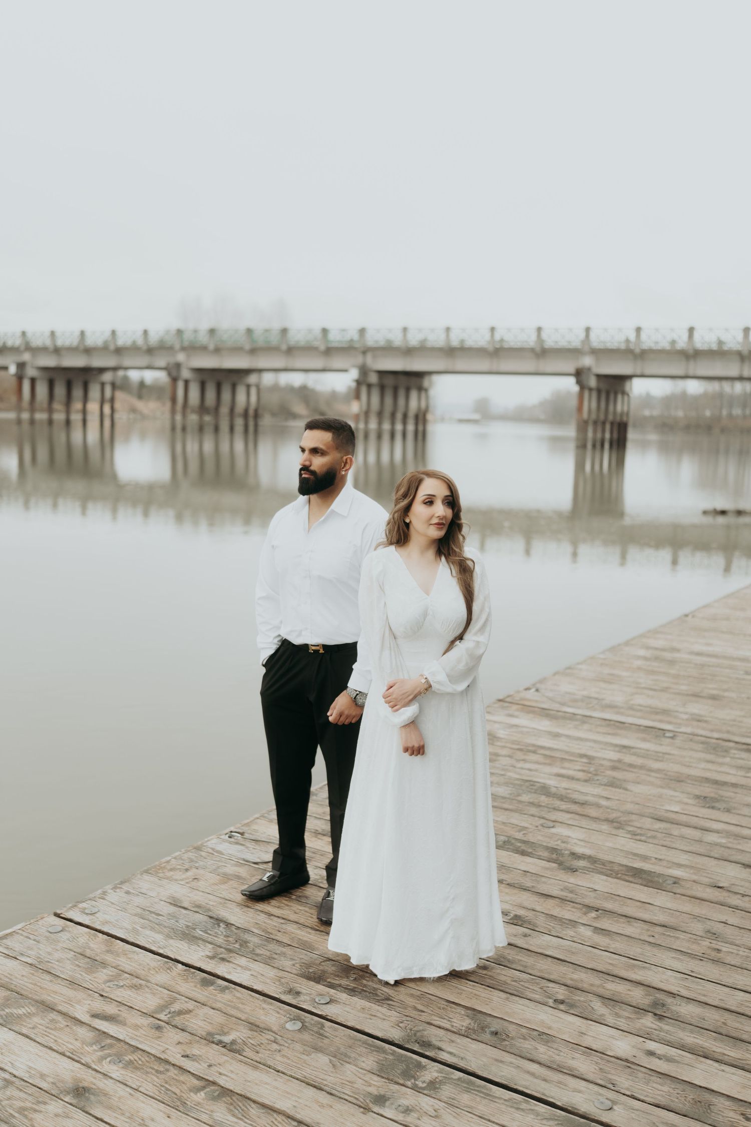 A couple in formal attire stands on a wooden boardwalk overlooking a misty lake and bridge.