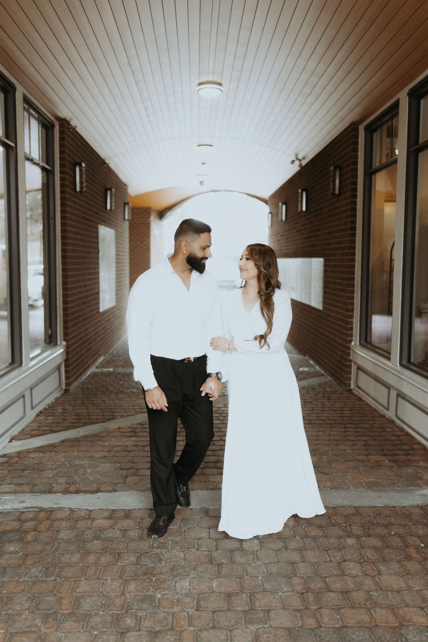 Playful couple poses in brick hallway wearing formal white dress and black outfit.