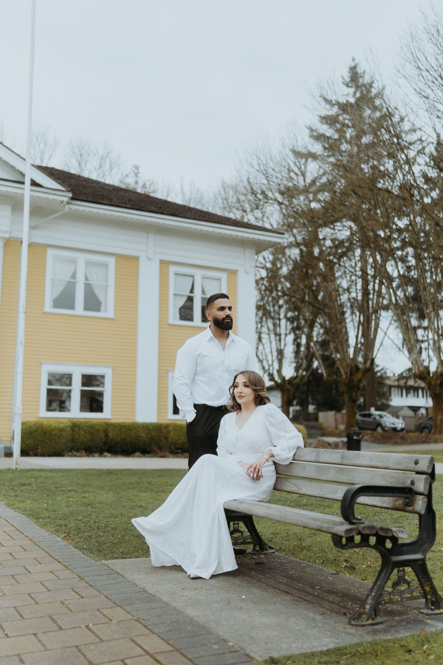 A couple poses for photos on a park bench outside a yellow colonial building.