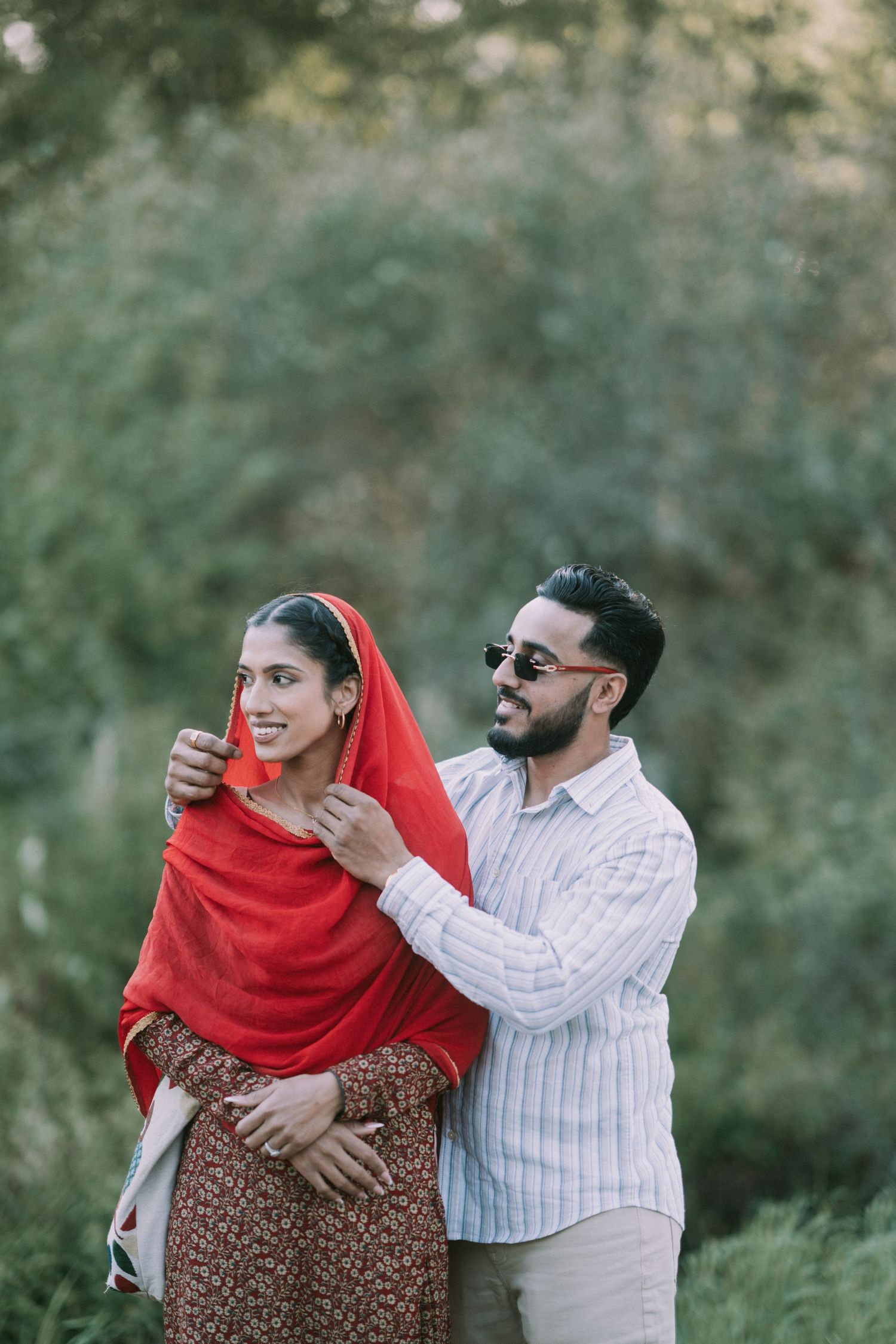 A couple shares a tender outdoor moment as she wears a red dupatta and floral dress while he dons a striped shirt.