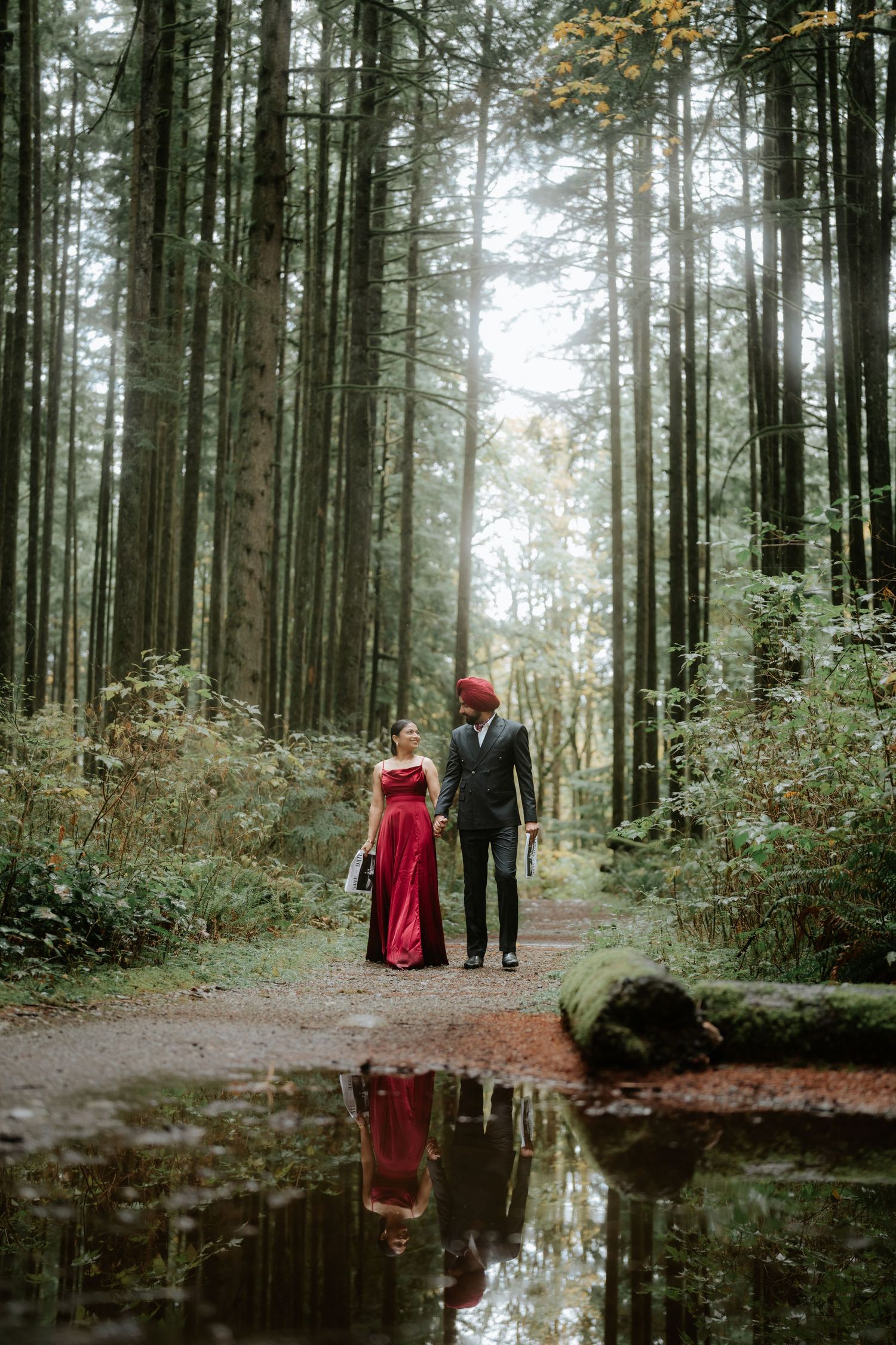 Couple walks along a reflective forest path surrounded by tall evergreen trees.