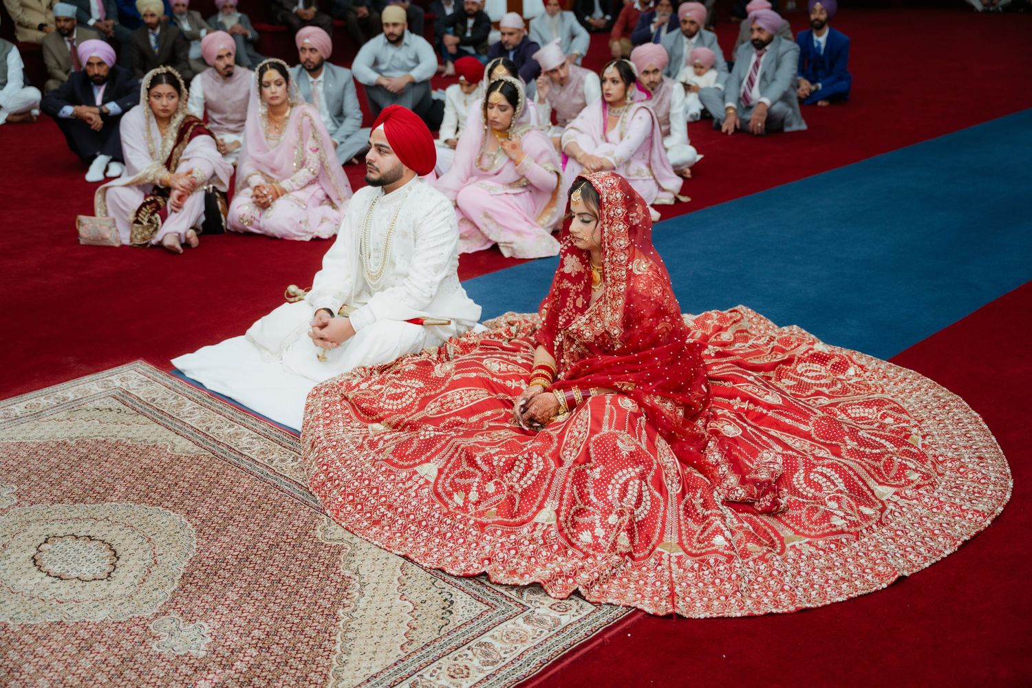 Wedding ceremony guests seated on floor with decorative red and blue carpet visible.