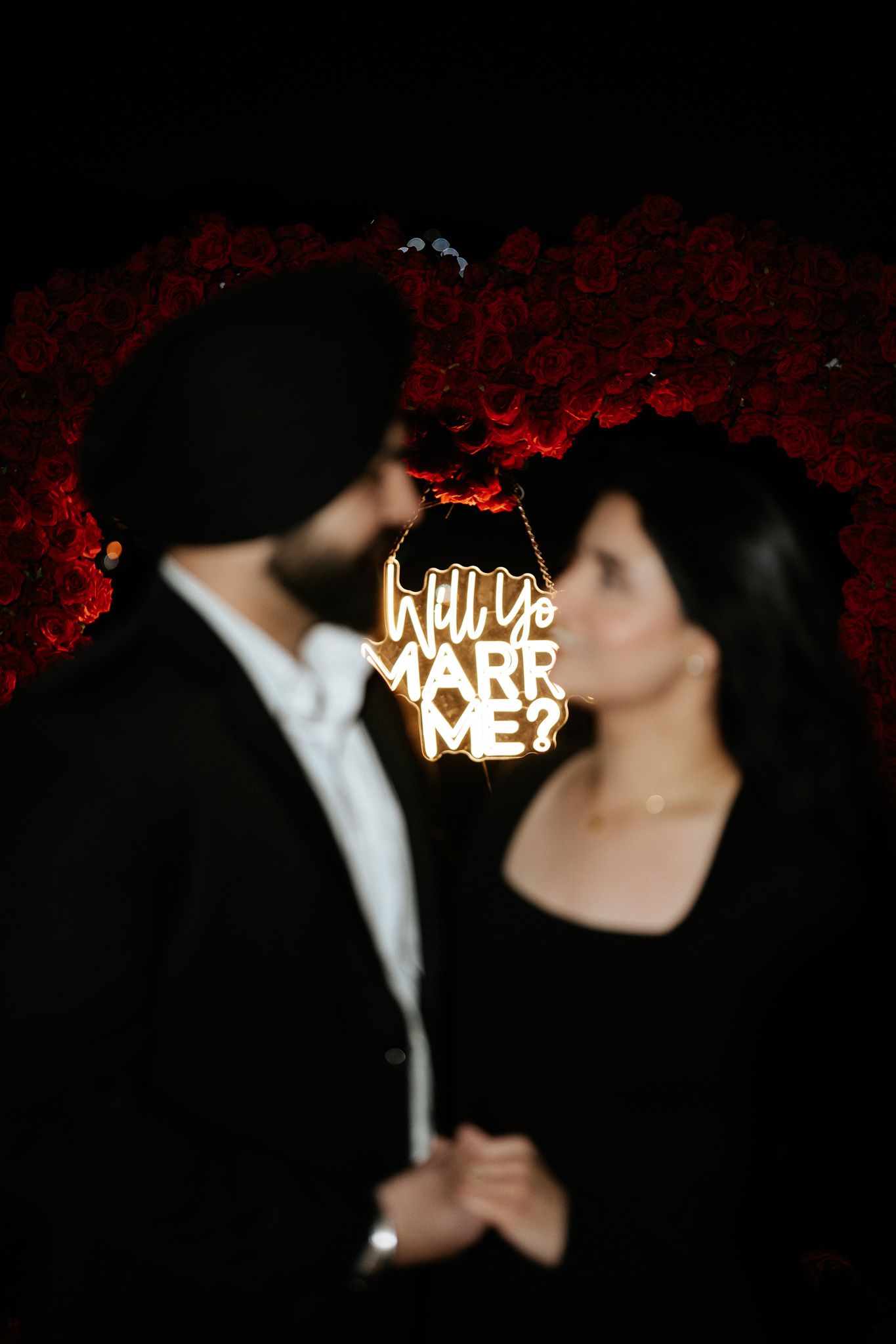 A romantic nighttime scene with a glowing 'Will You Marry Me' sign and red floral decorations.