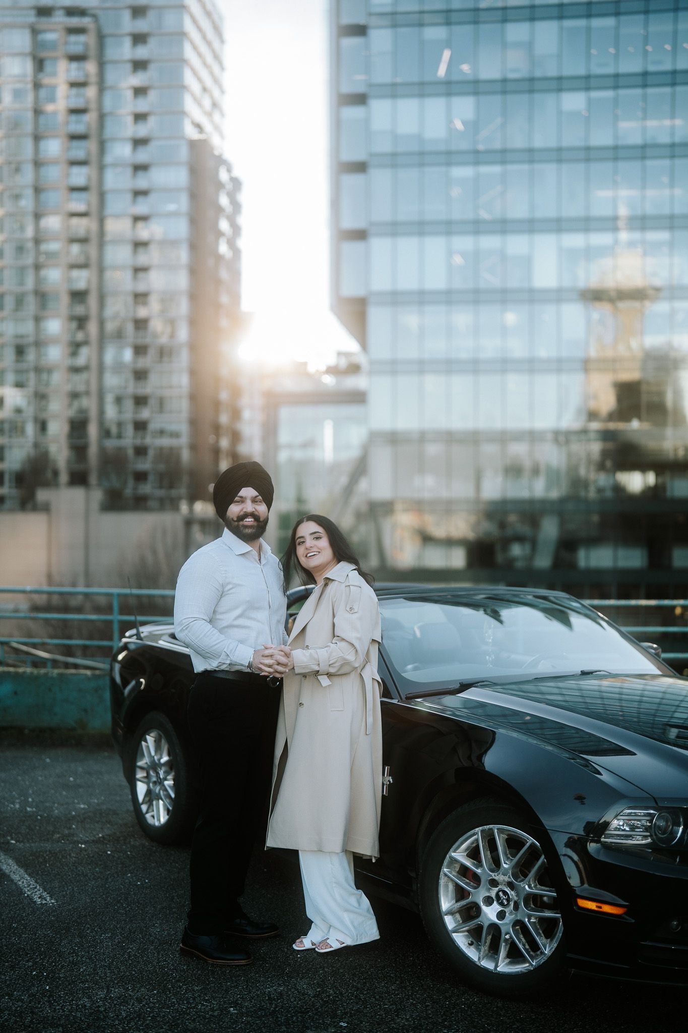 A couple poses next to a black Ford Mustang on a rooftop parking area with modern glass buildings in the background at sunset.