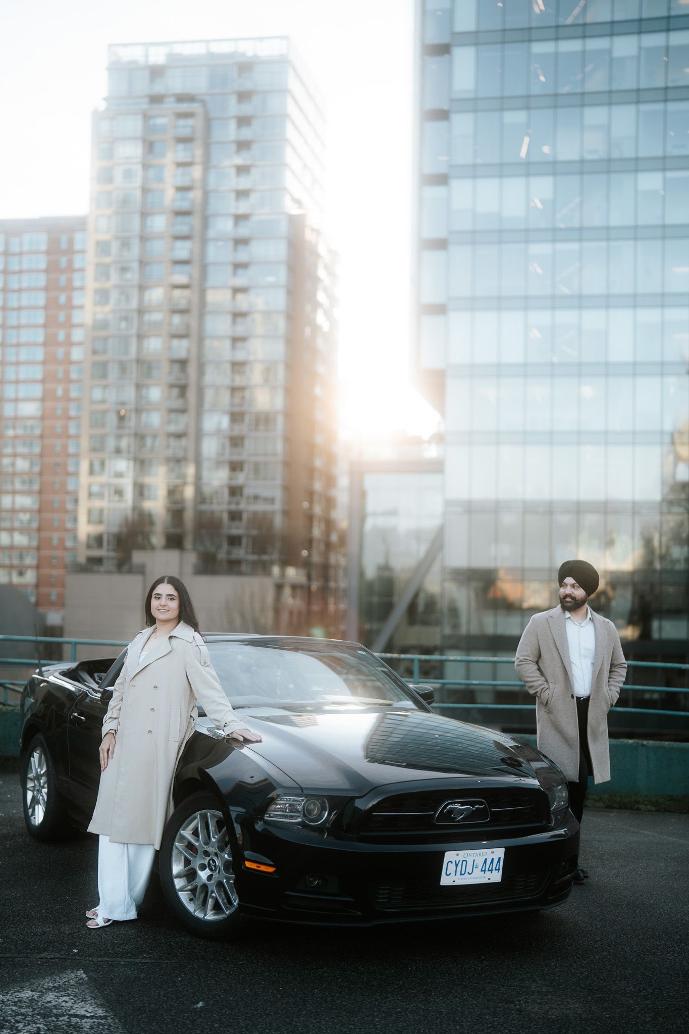 A black Ford Mustang parked in front of a modern glass building at sunset with a person posing beside it.