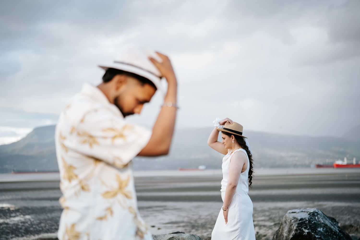 Couple in straw hats posing outdoors near ocean with cloudy sky, man in floral shirt and woman in white dress.
