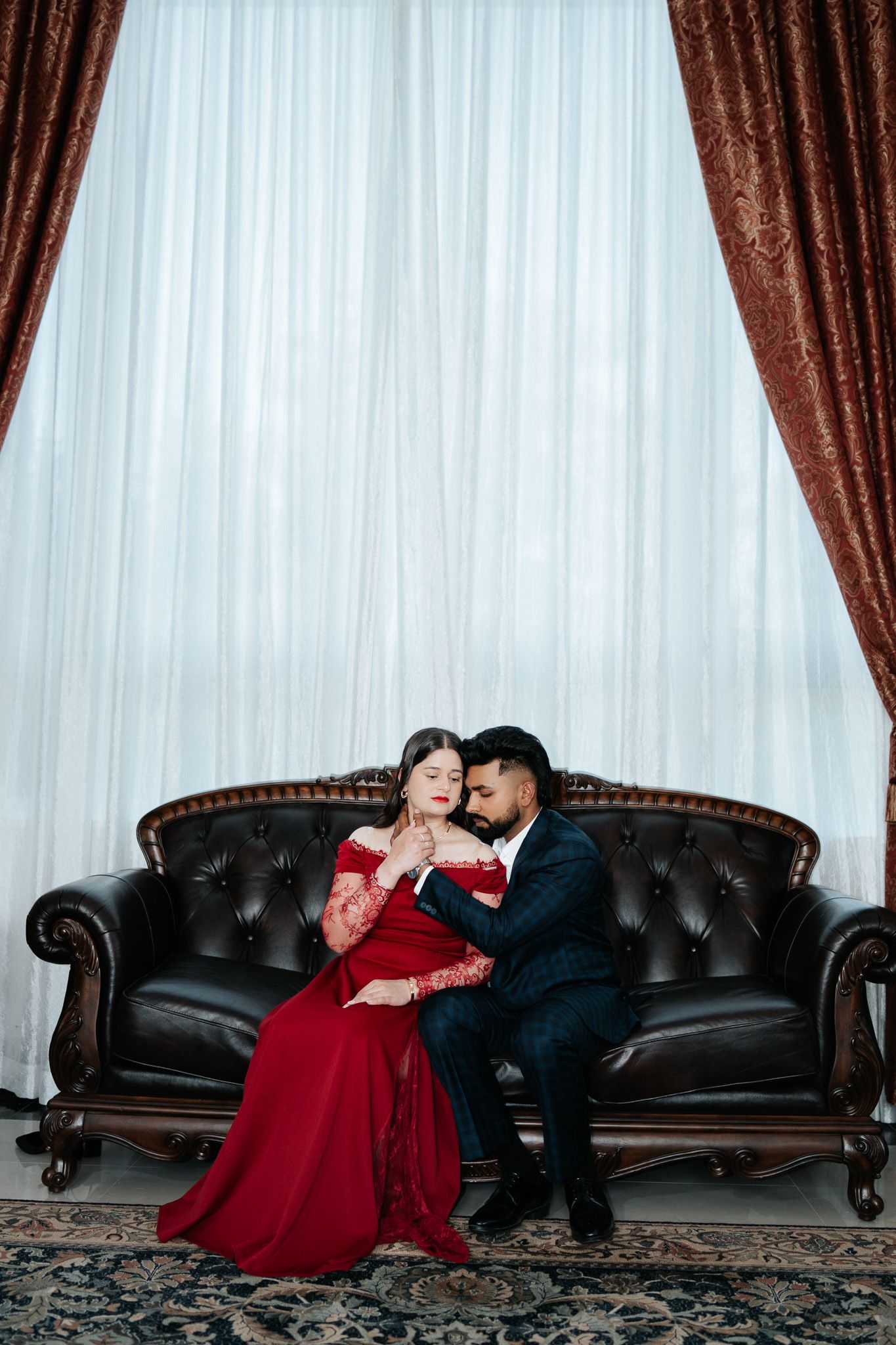 Elegant couple in red dress and dark suit sitting intimately on a tufted leather sofa with red curtains.