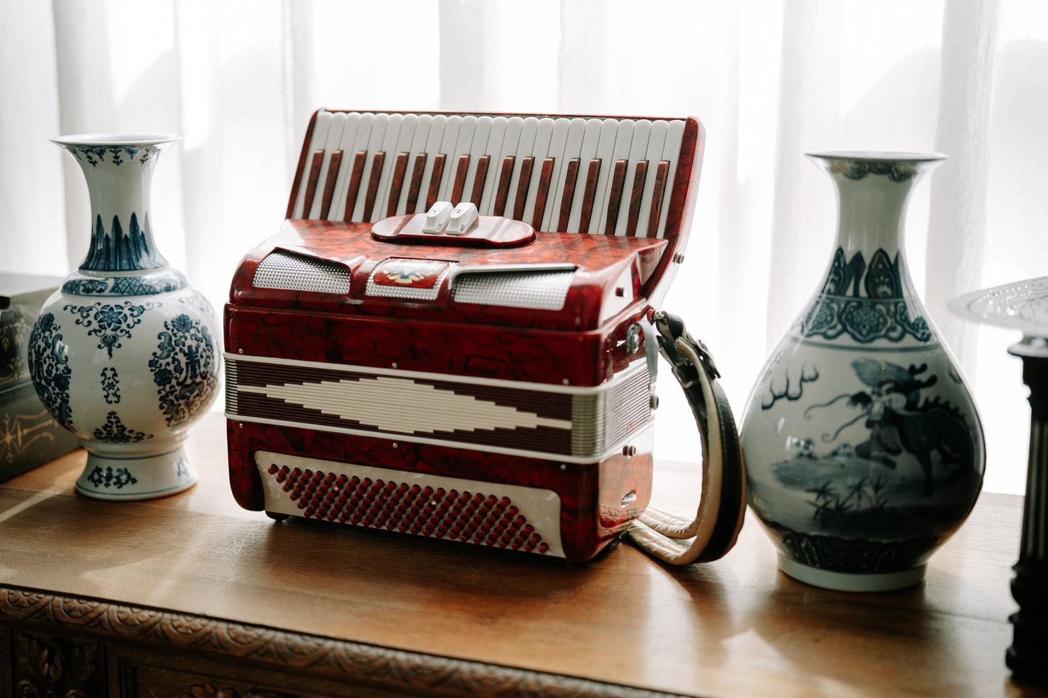 Red accordion on wooden table flanked by blue and white Chinese porcelain vases near a window.