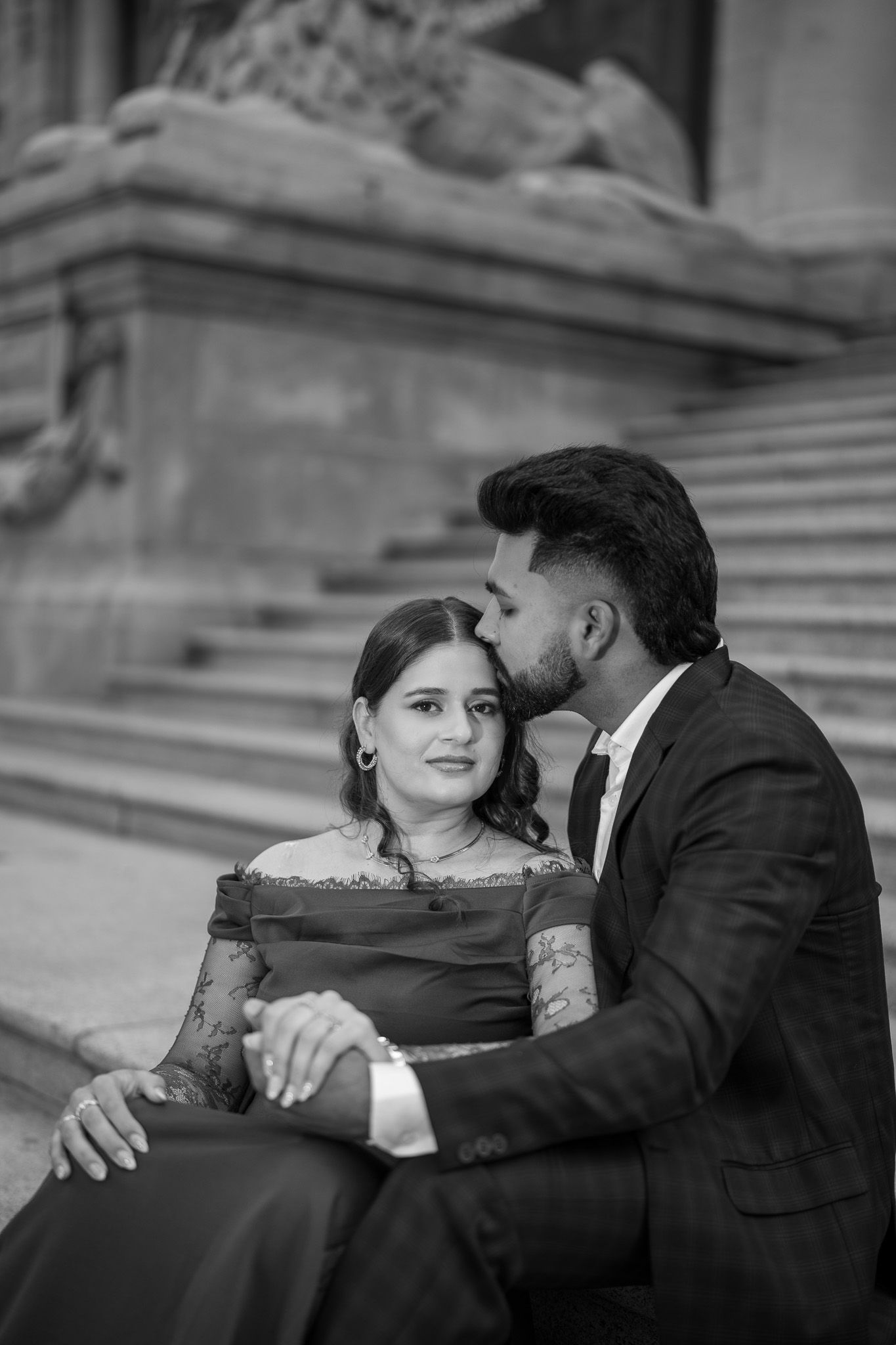 Black and white photo of a couple sitting on steps, man kissing woman on cheek, both dressed in formal attire.