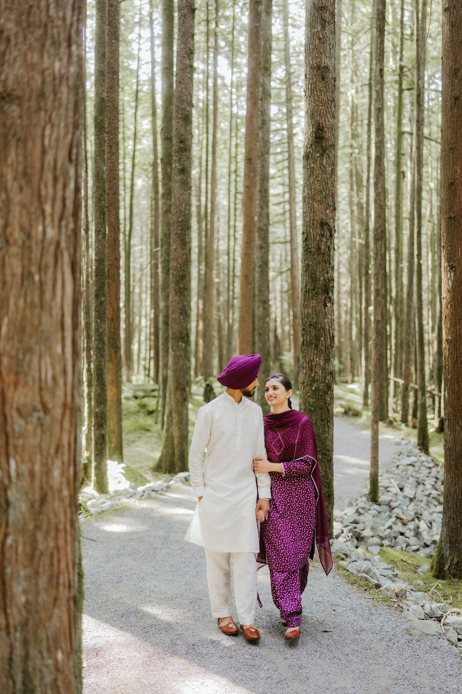 Sikh couple walking hand-in-hand through a tall forest, wearing traditional purple and white attire.