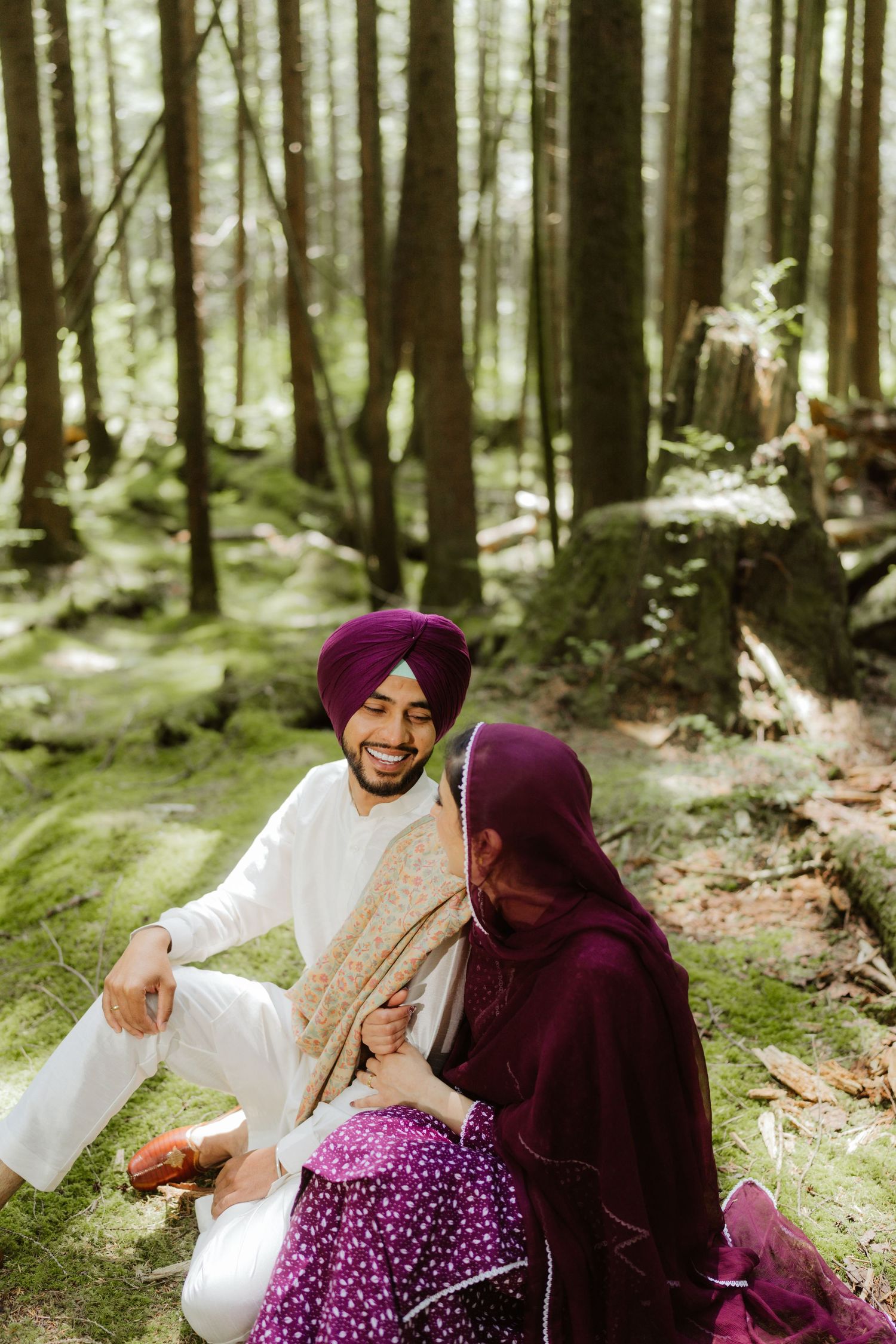 Smiling Sikh man in white kurta and purple turban sitting with woman in purple salwar in a lush green forest.