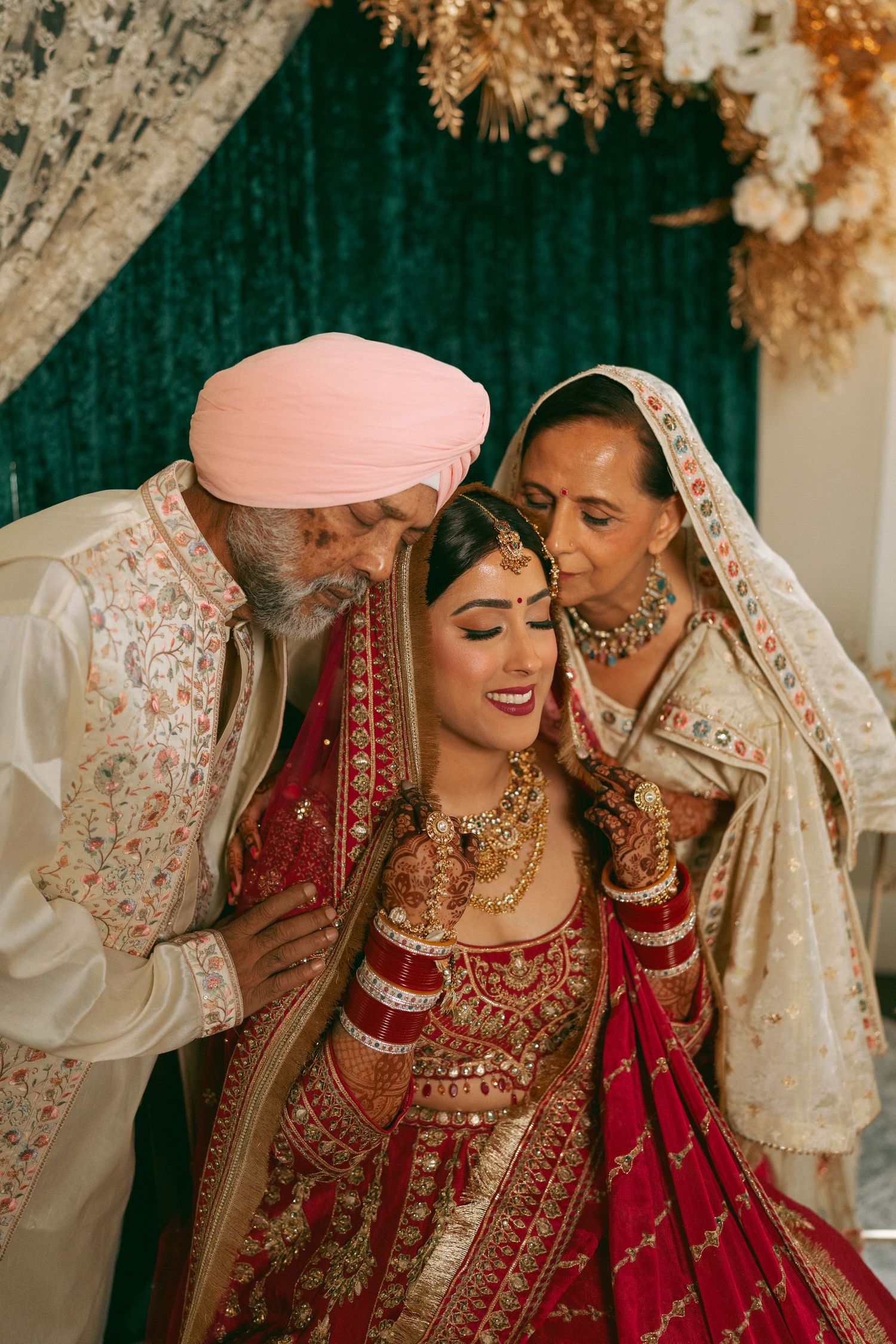 Sikh bride in red lehenga with gold jewelry receives blessings from parents during traditional wedding ceremony.
