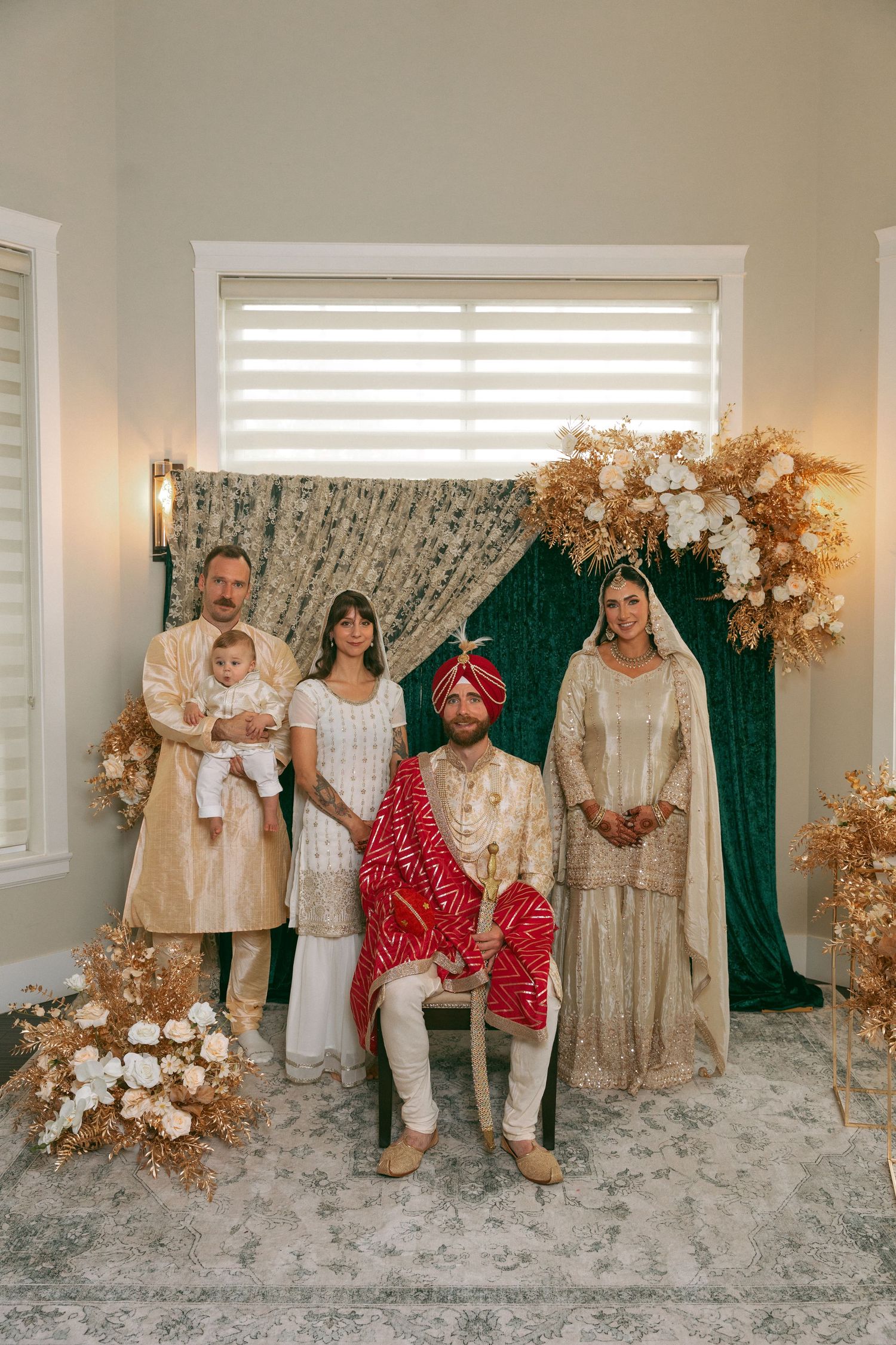 Family poses in traditional South Asian wedding attire before elegant floral backdrop with gold and white blooms.