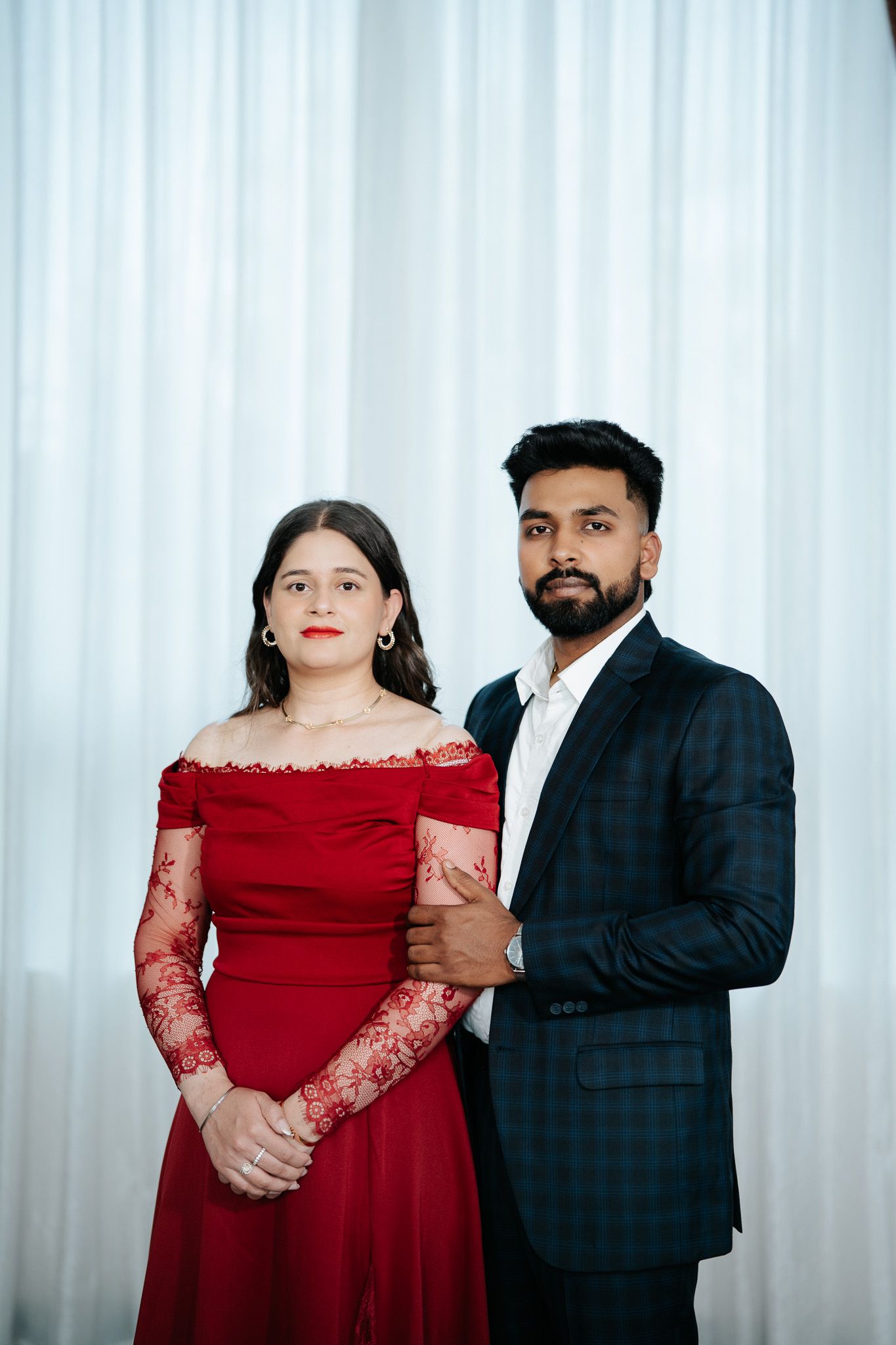 Indian couple posing together, woman in red lace off-shoulder gown and man in dark plaid suit.