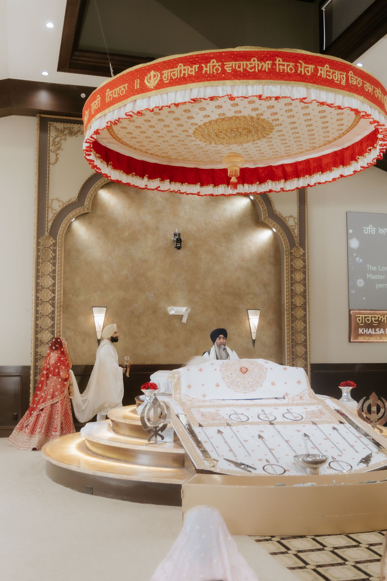 Sikh wedding ceremony at a Gurdwara with bride in red attire circling the Guru Granth Sahib under a decorative canopy.