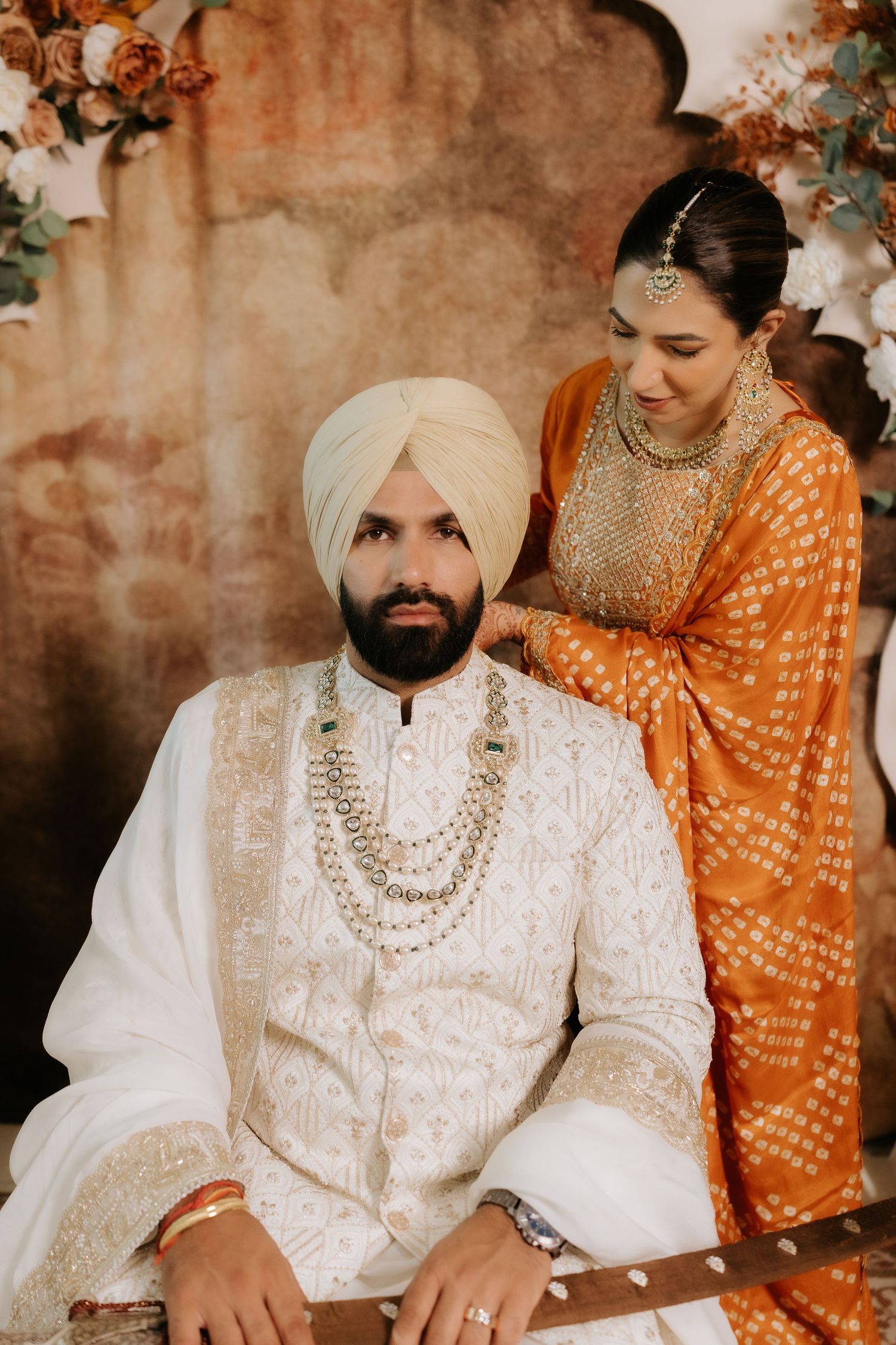 Sikh couple in traditional wedding attire, groom in white sherwani and turban, bride in orange embroidered suit.