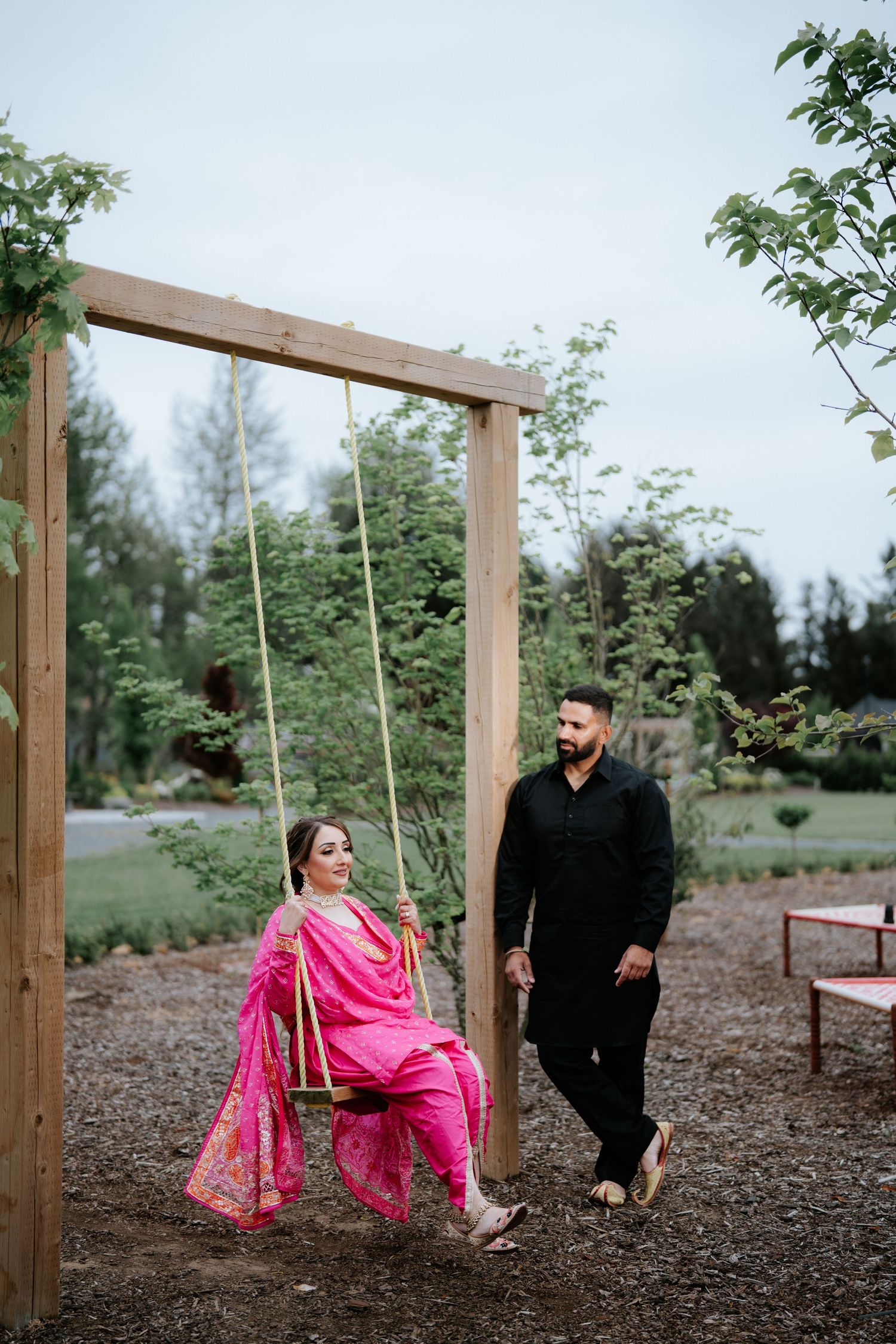 A woman in pink salwar kameez sits on a wooden swing while a man in black stands beside her in a lush garden.