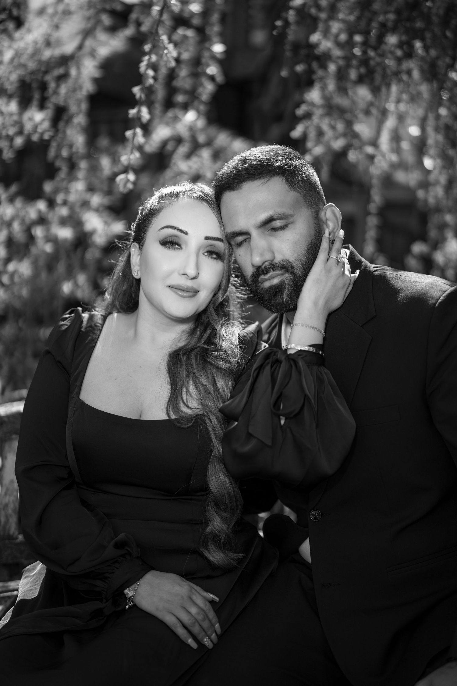 Black and white portrait of a couple in elegant black attire, posing intimately against a lush garden backdrop.