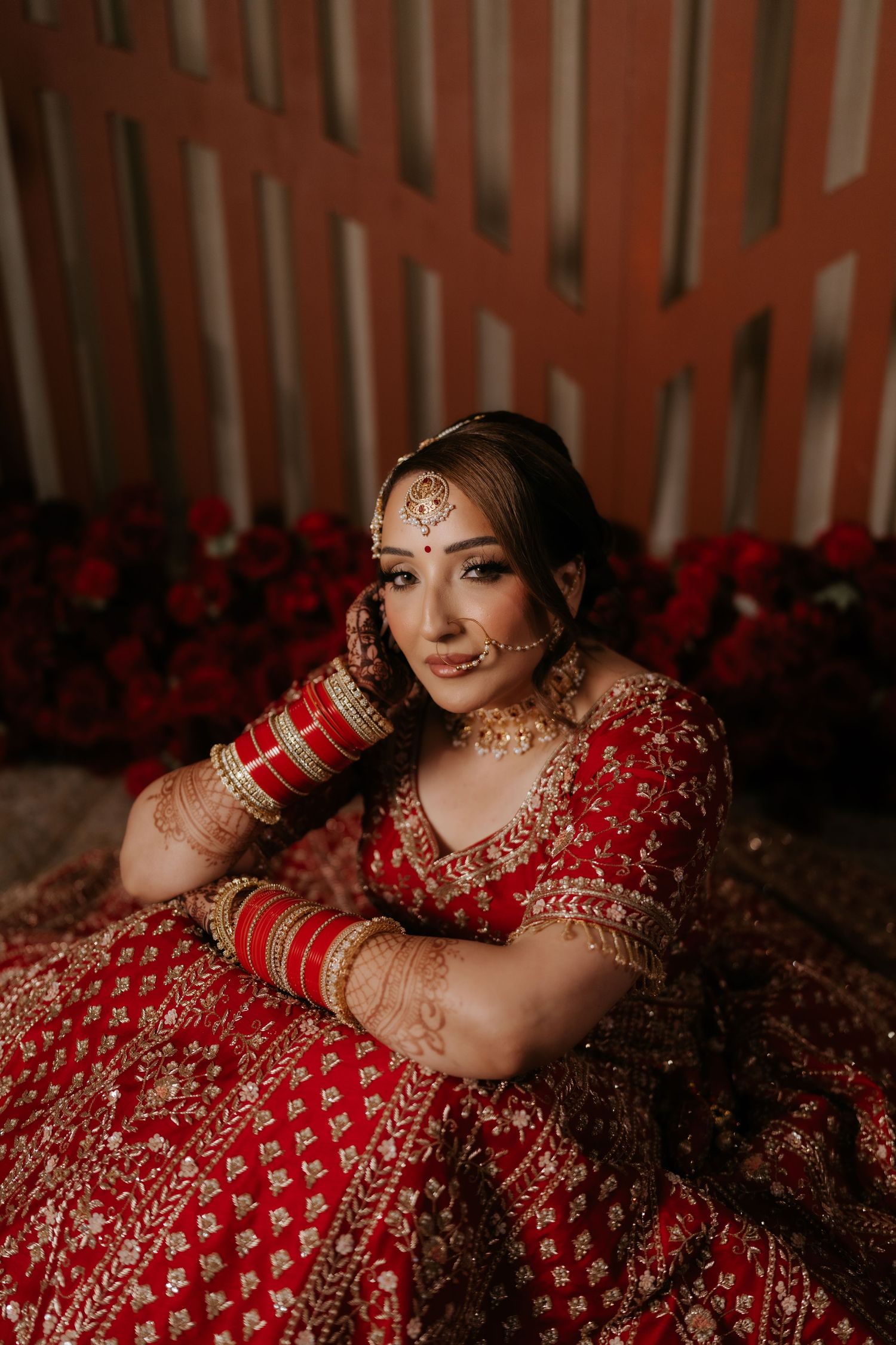 Indian bride in red lehenga with gold embroidery, bridal jewelry, and mehndi, posing gracefully against a floral backdrop.