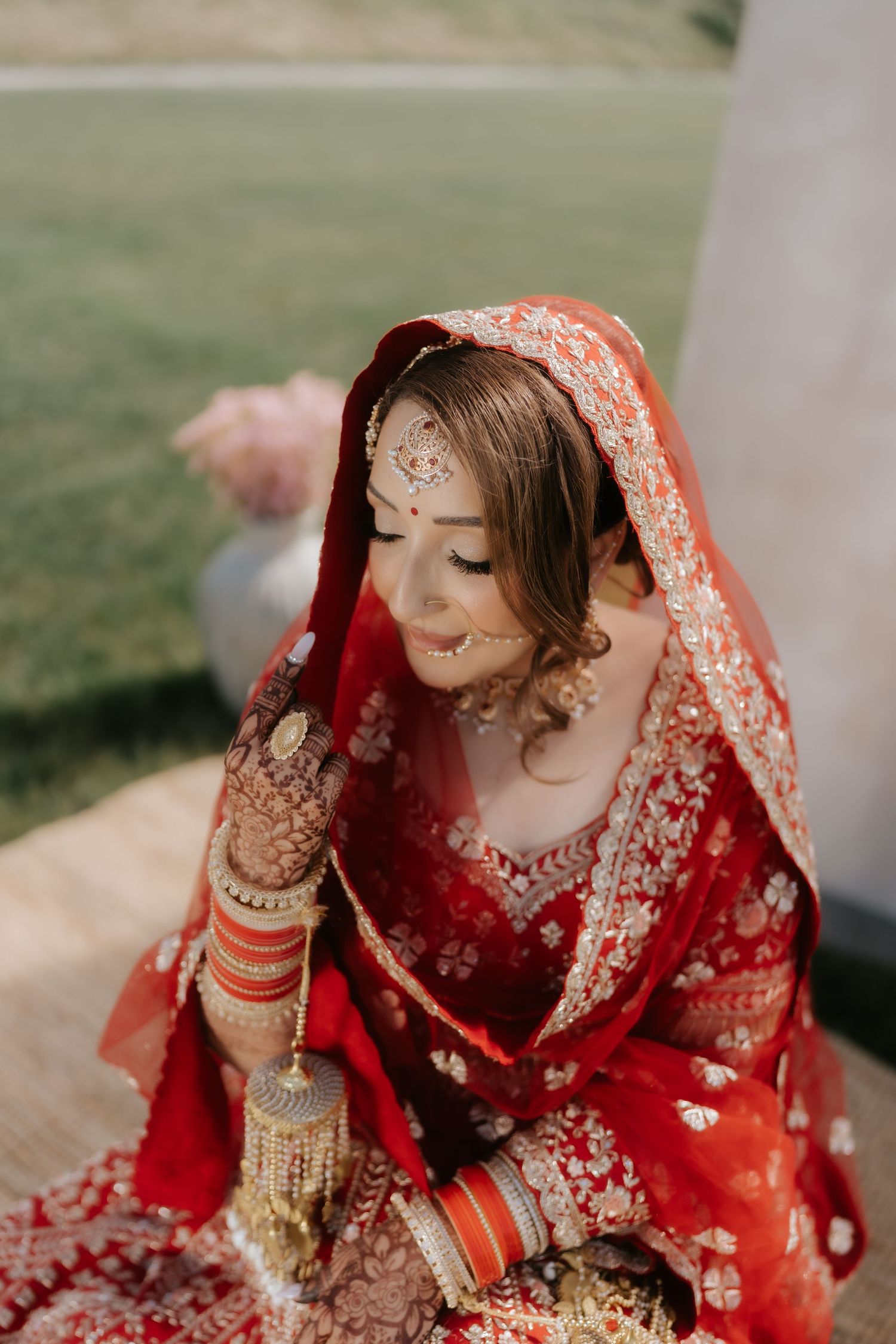 Indian bride in red lehenga with gold embroidery, wearing traditional jewelry and dupatta, looking down gracefully.