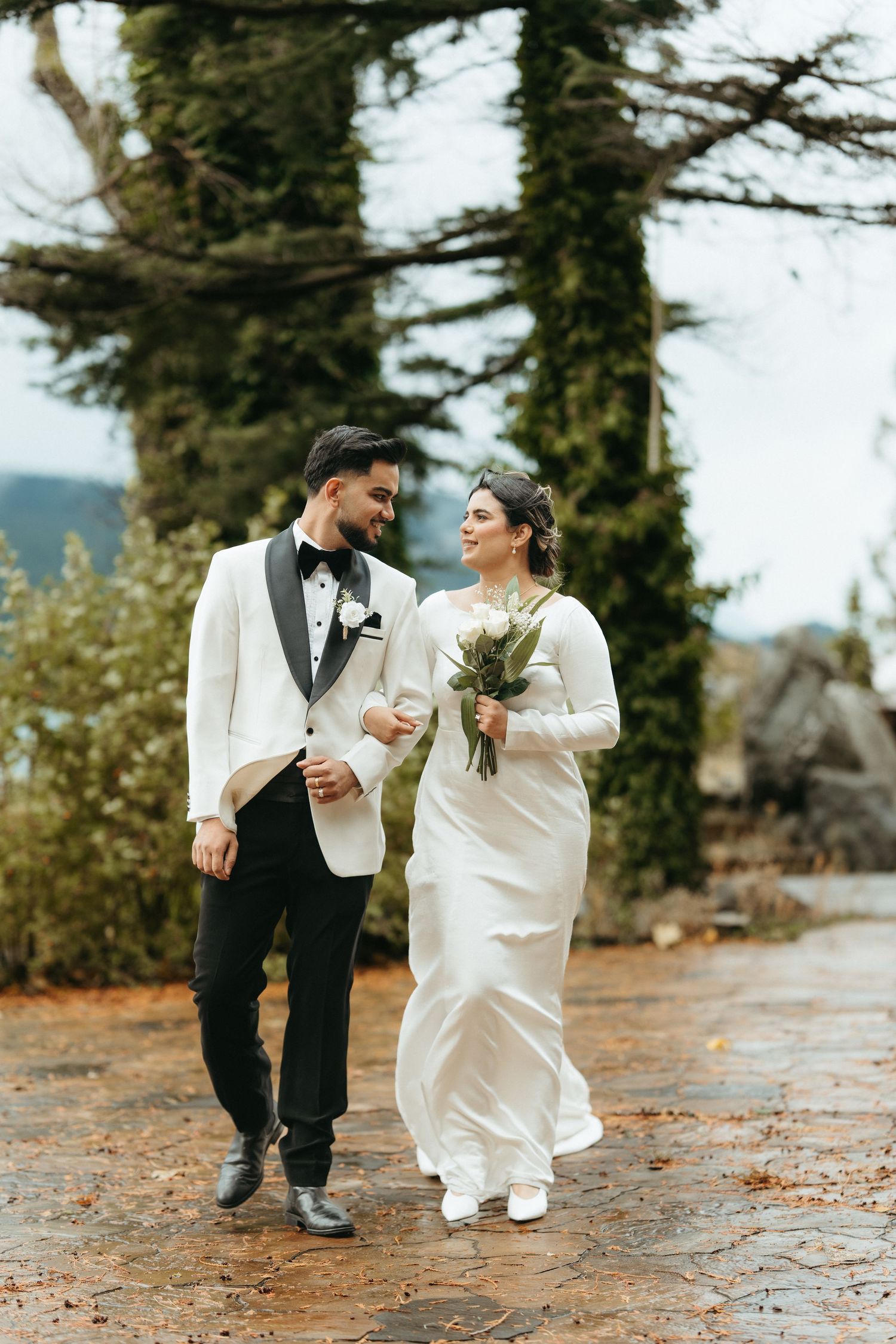 A joyful couple in wedding attire share a laugh while walking through a forest path on their special day.