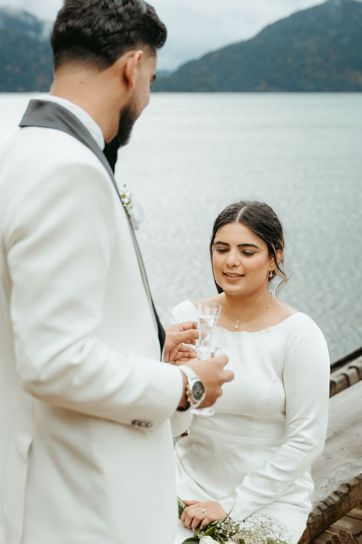 A couple in white wedding attire shares an intimate moment by a misty lake with mountains in the background.