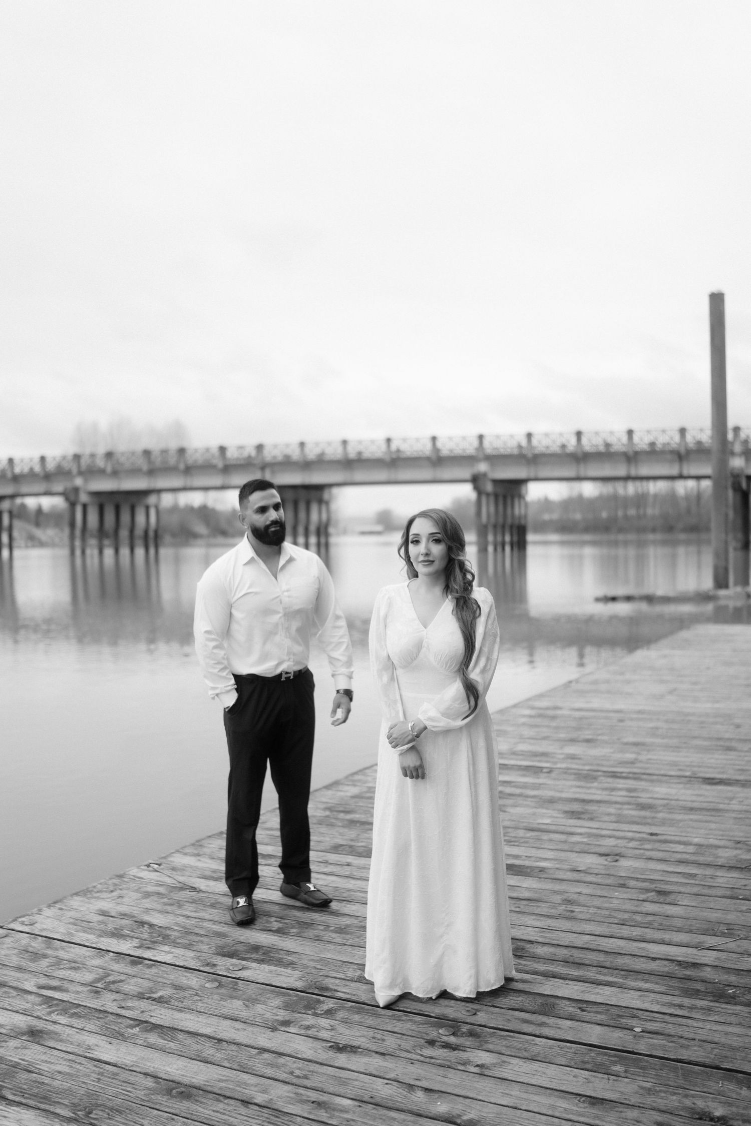 A moody black and white photo of a couple on a wooden dock with a bridge in the background.