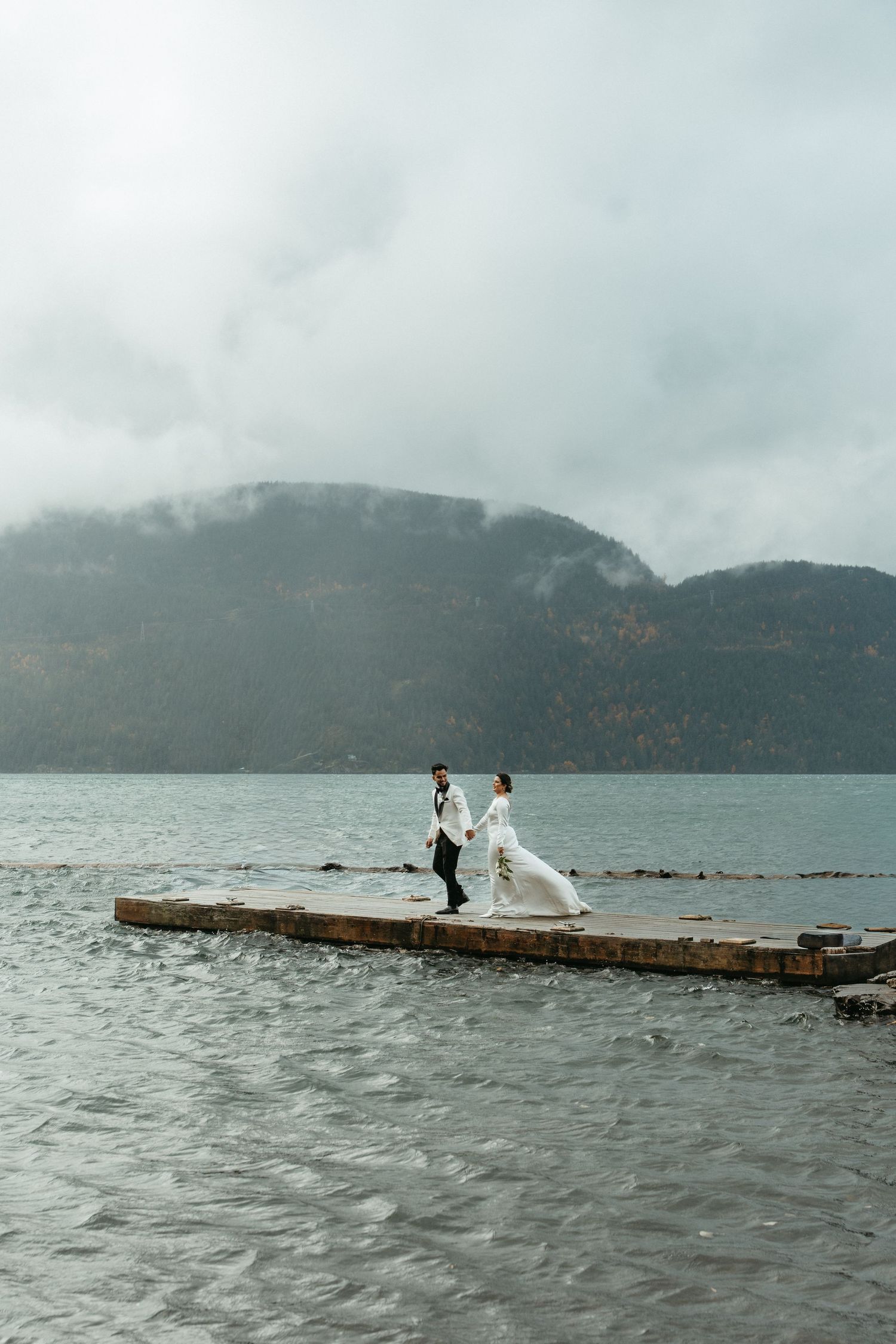 Two people stand on a wooden dock overlooking a misty mountain lake on an overcast day.