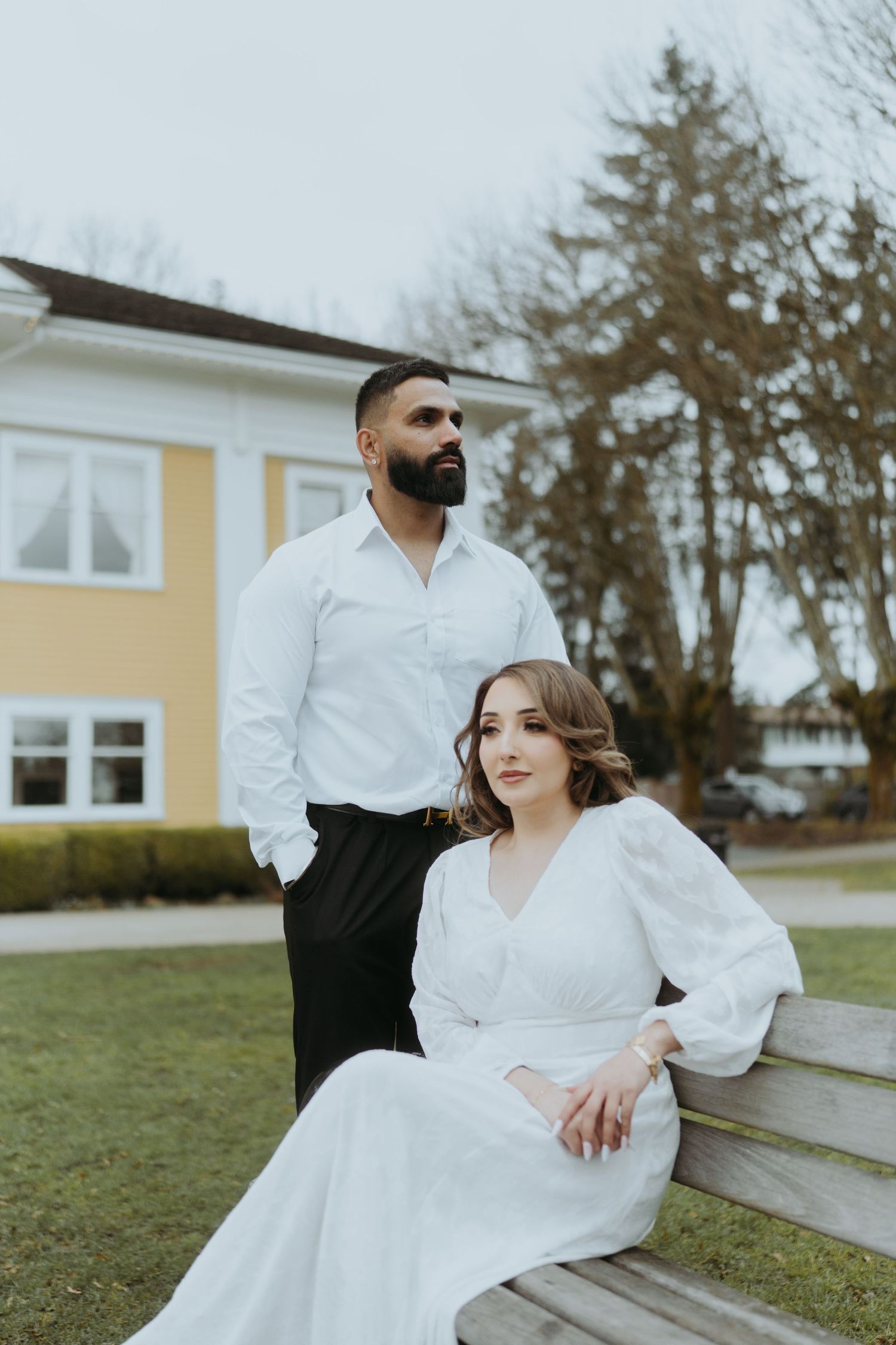 A couple poses together near a park bench with evergreen trees in the background.
