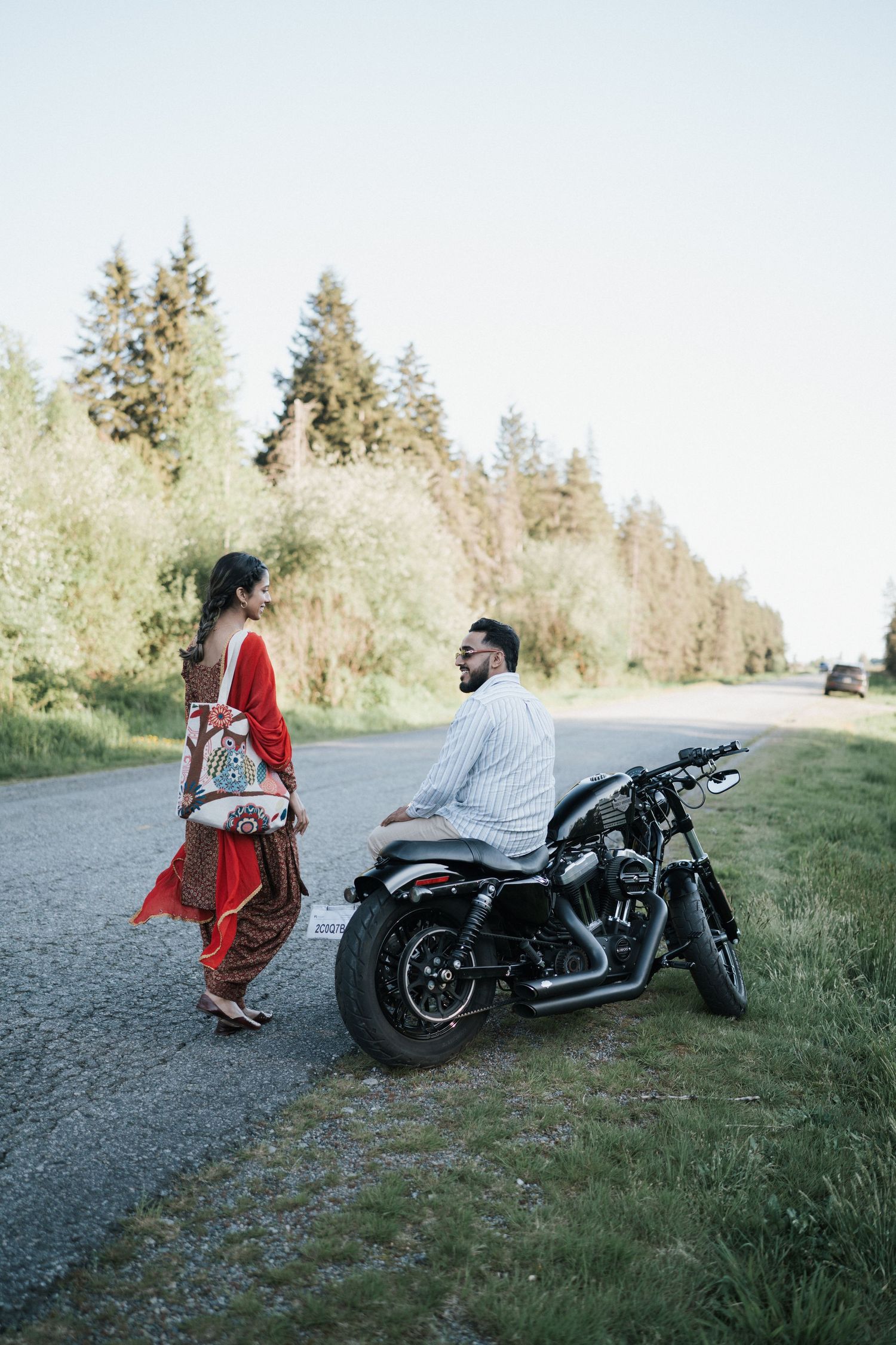 Two people share a moment on a rural road with a motorcycle, surrounded by evergreen trees and grass.