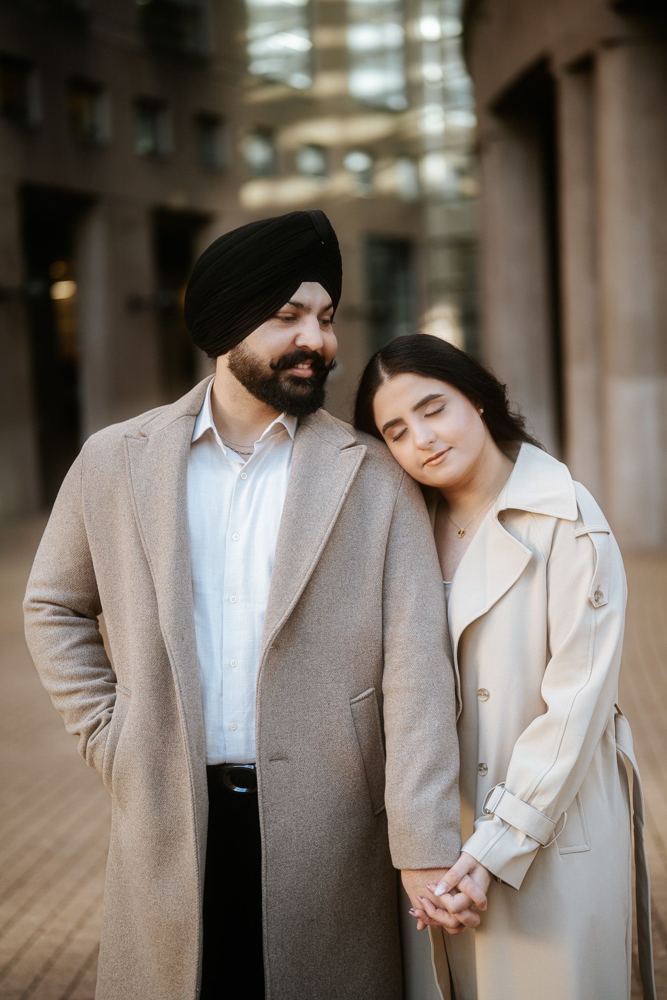 A couple in matching beige coats hold hands while standing together in a modern indoor setting.