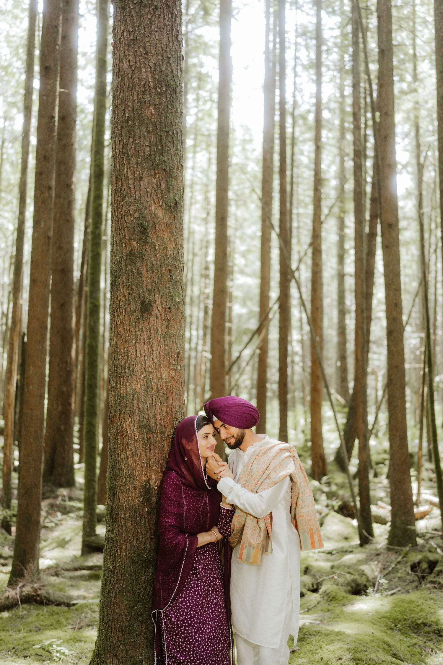 Sikh couple in traditional attire share a romantic moment leaning against a tree in a sunlit forest.