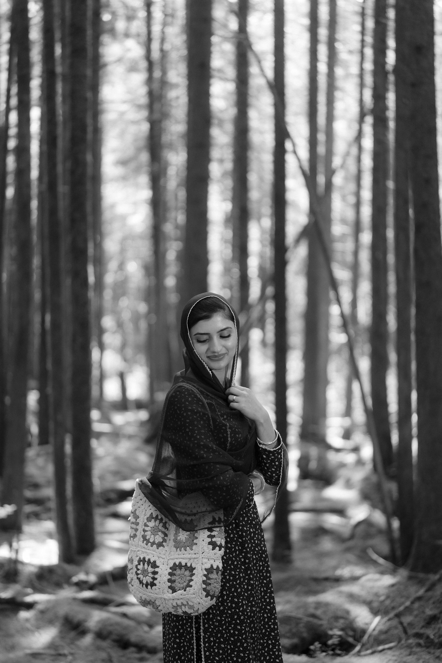 Black and white portrait of a South Asian woman in a headscarf holding a crochet bag in a tall pine forest.