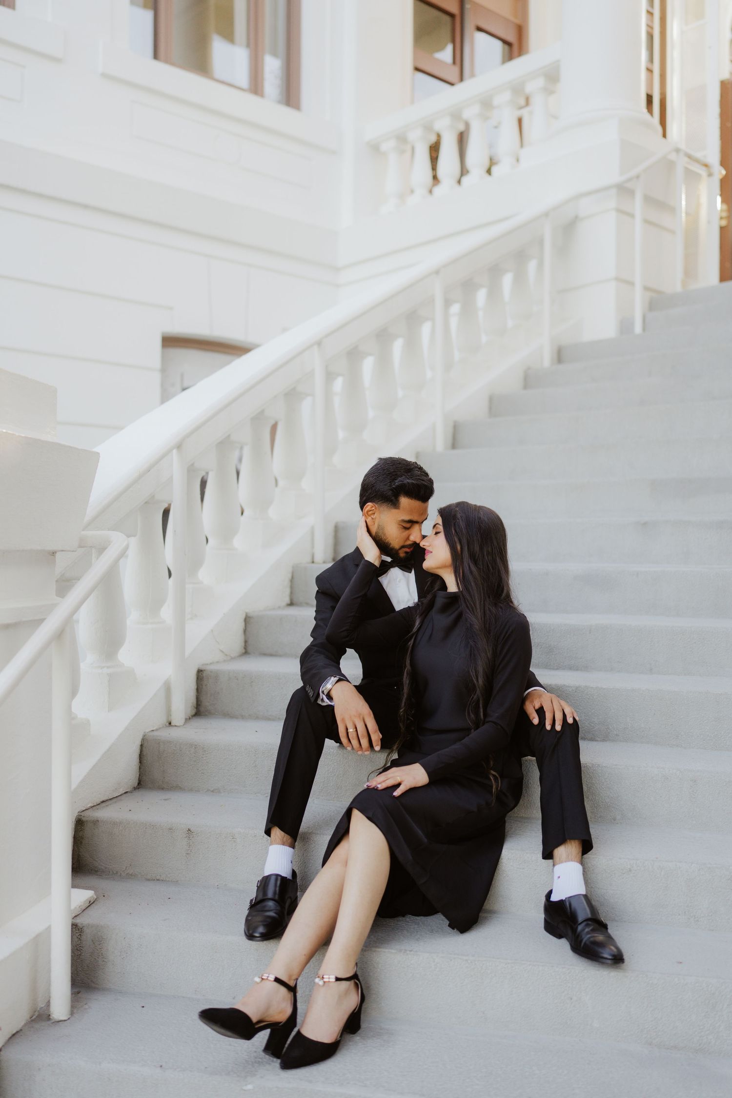 Couple in matching black outfits sharing a romantic moment on elegant white marble stairs of a classical building.