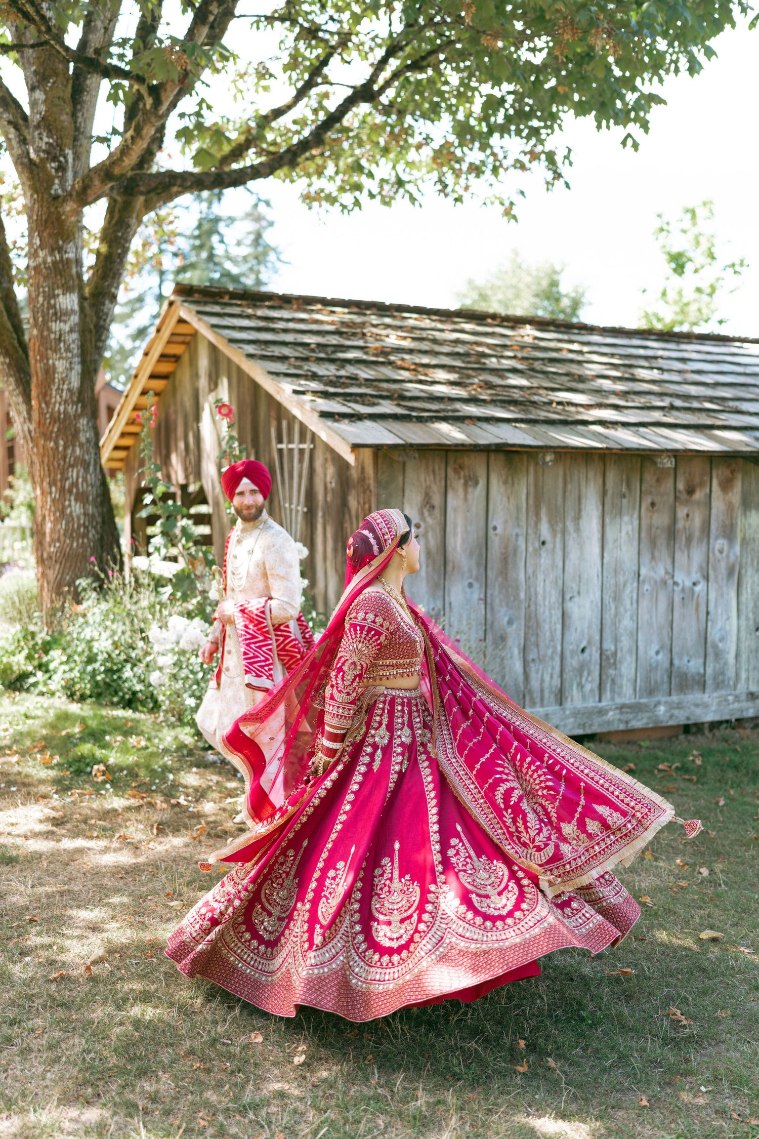 Indian bride twirling in vibrant red lehenga with gold embroidery outdoors, groom in red turban standing nearby.