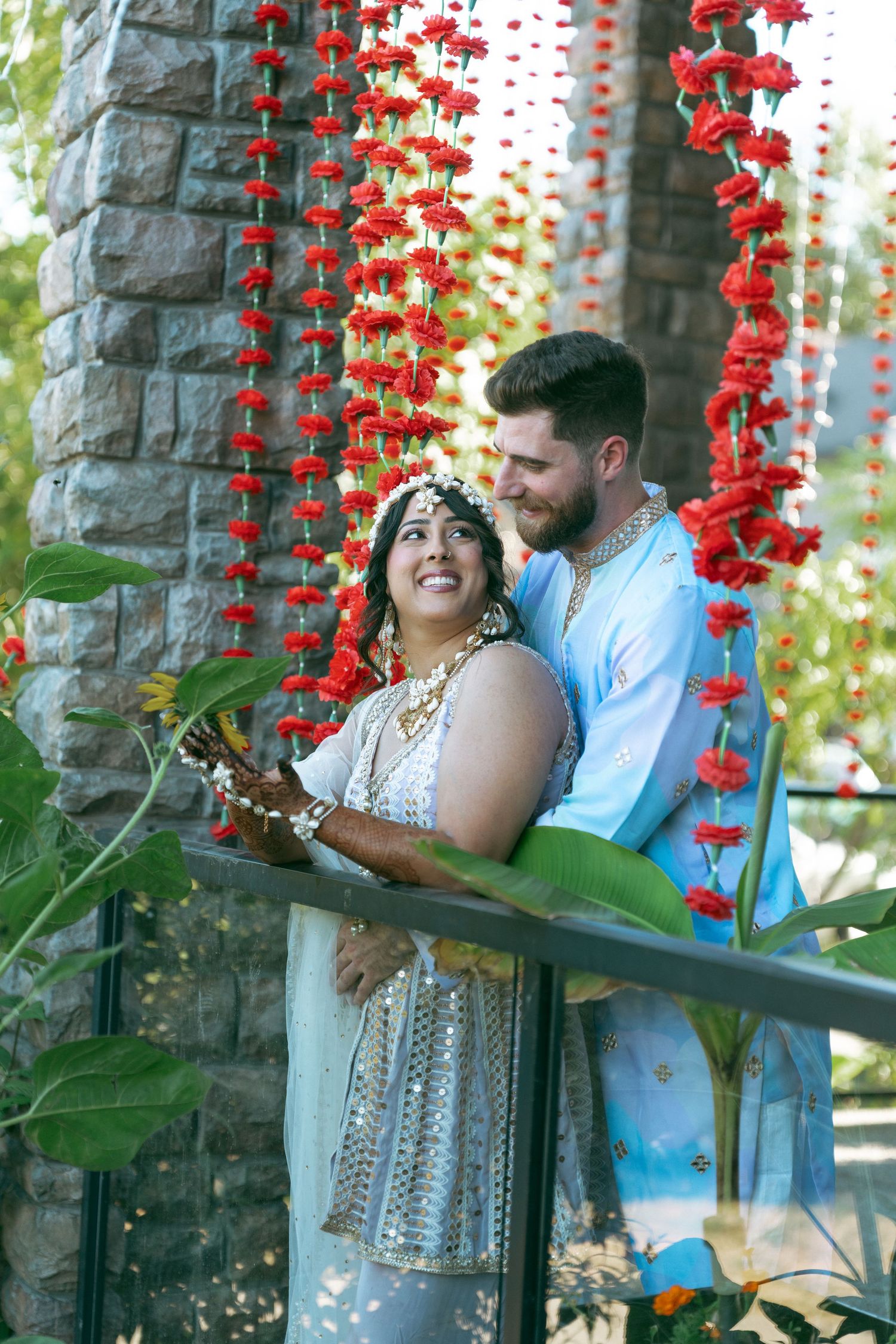 Couple in traditional Indian attire smiling together under red floral garlands at an outdoor wedding ceremony.