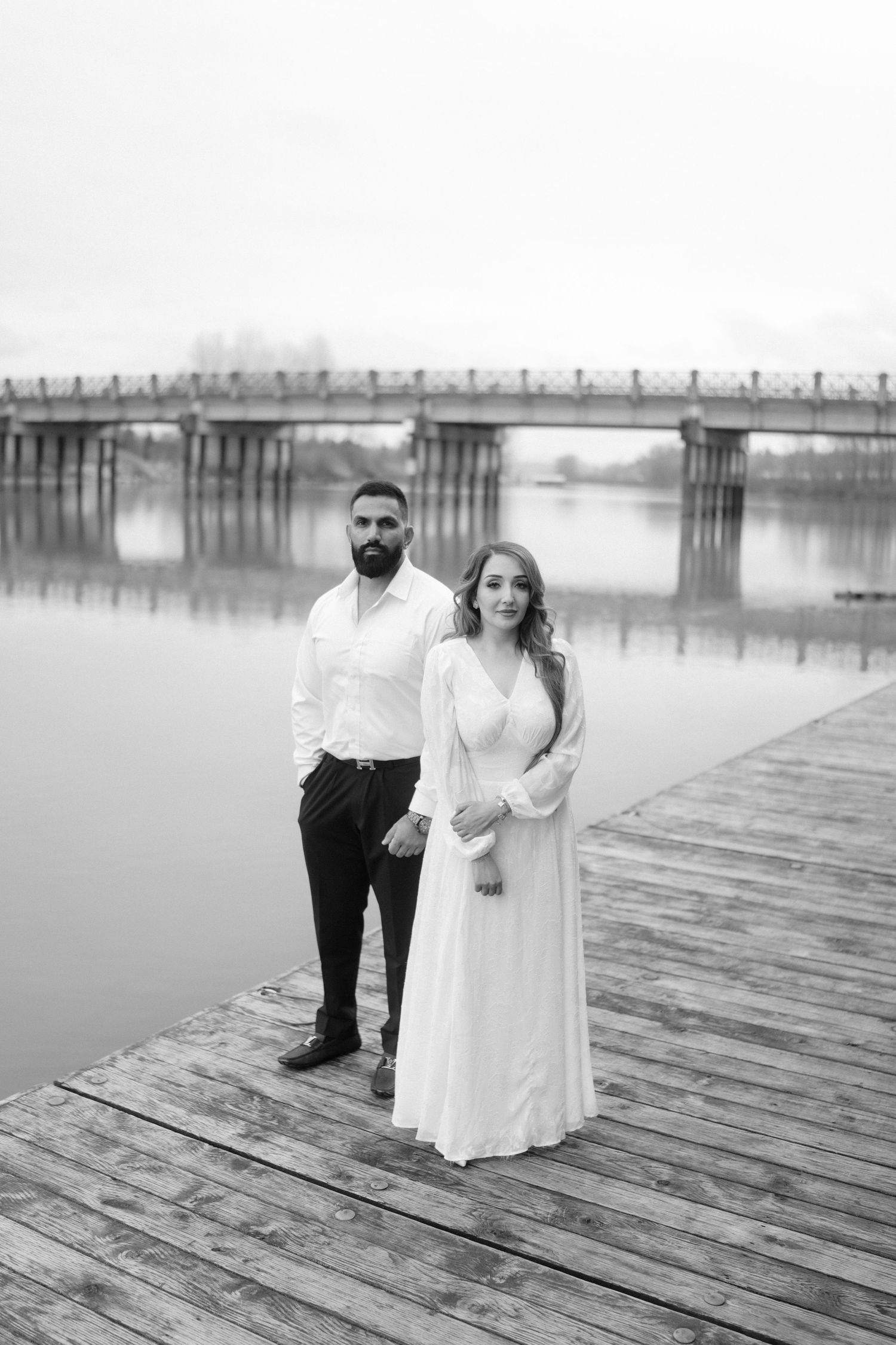A black and white photo of a couple standing on a wooden pier with a bridge in the background.