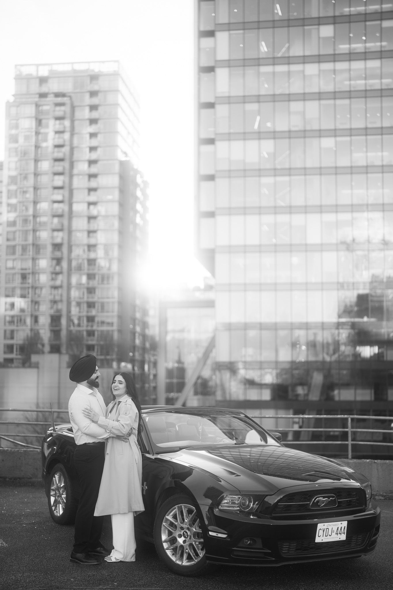 A black luxury car parked in front of a modern glass building with a couple standing nearby at sunset.