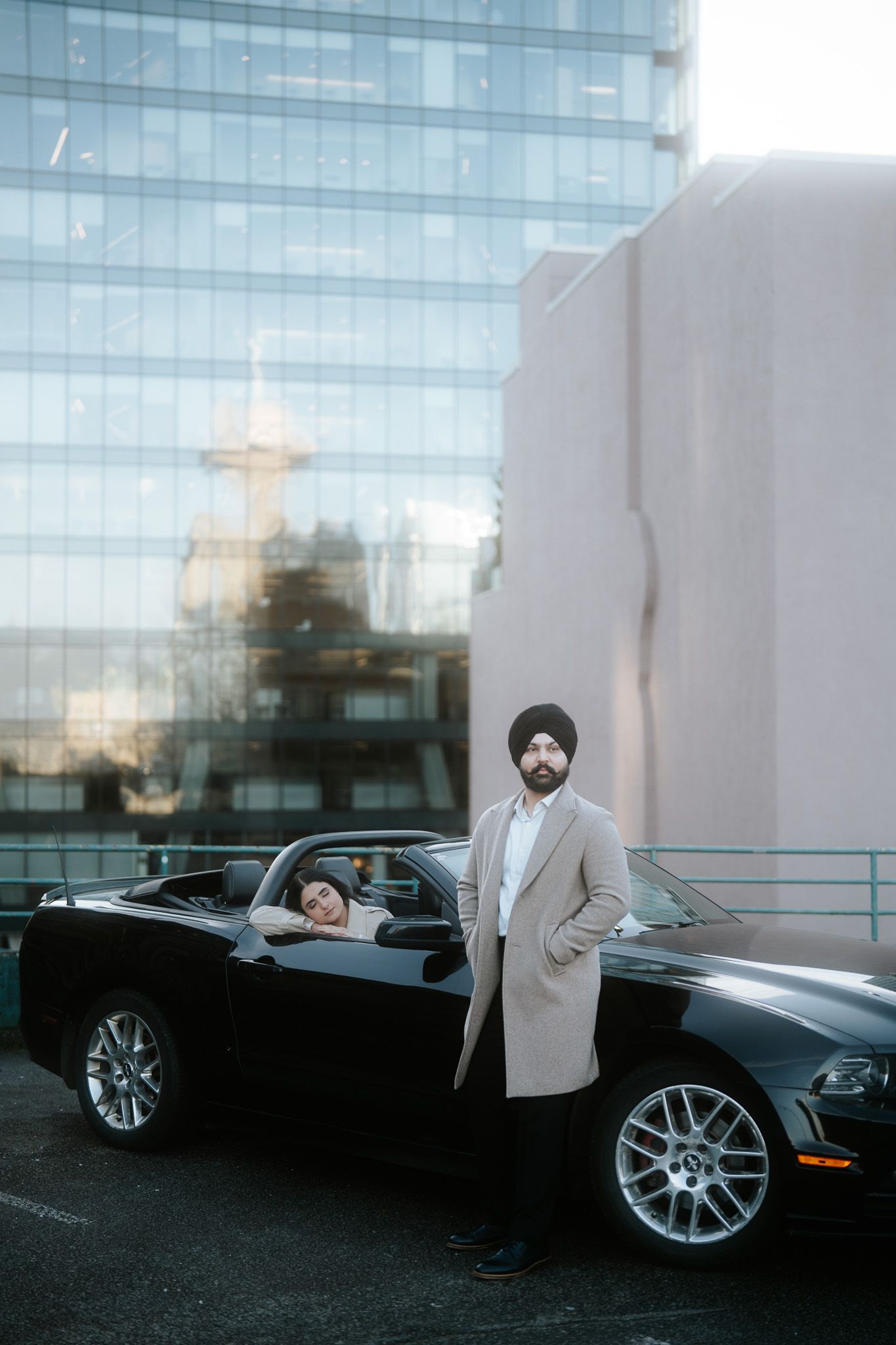 Luxury black car with chrome wheels parked against modern glass building exterior in evening light.