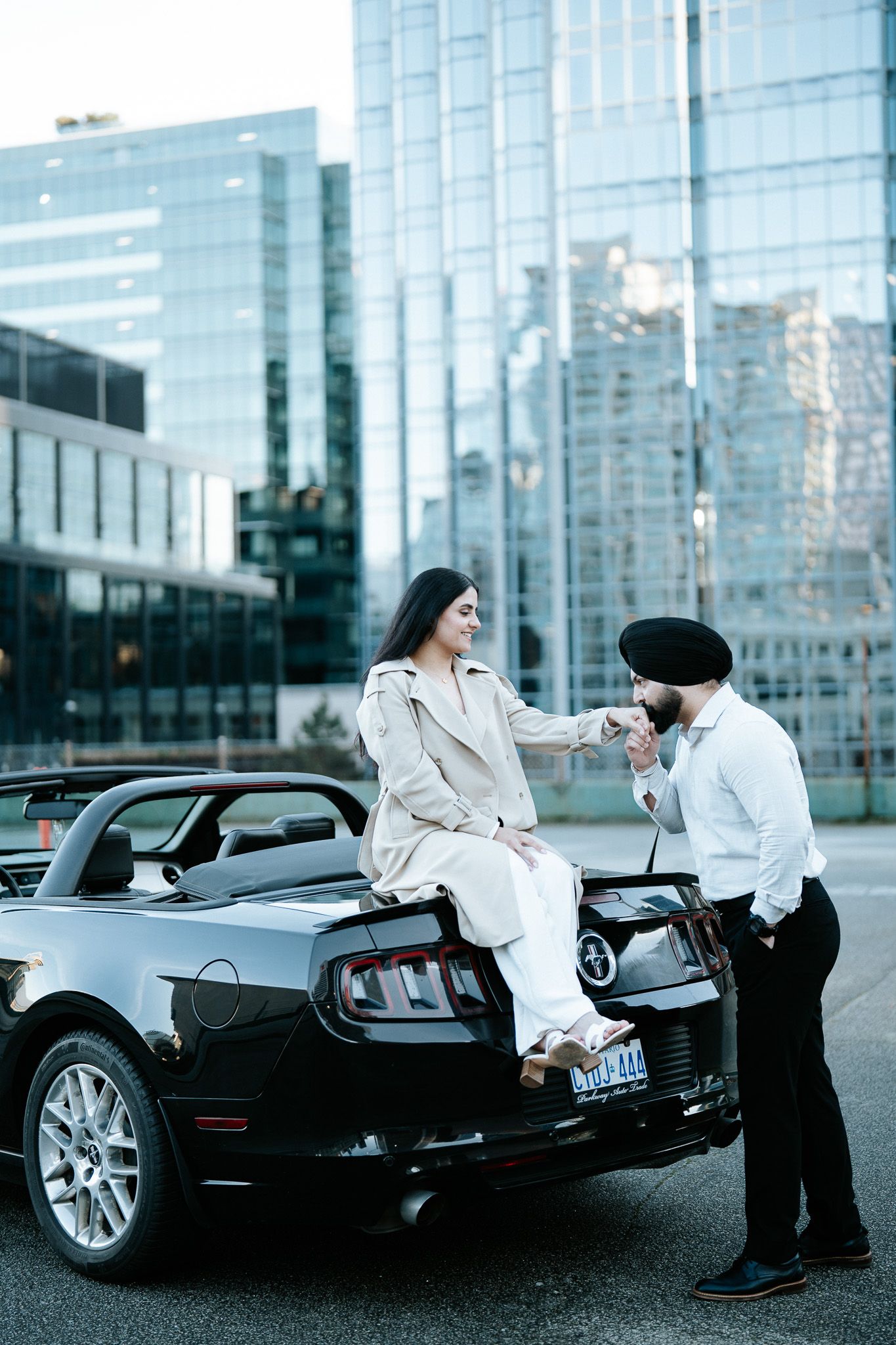 A stylish couple poses with a black luxury sports car against a modern glass building backdrop.