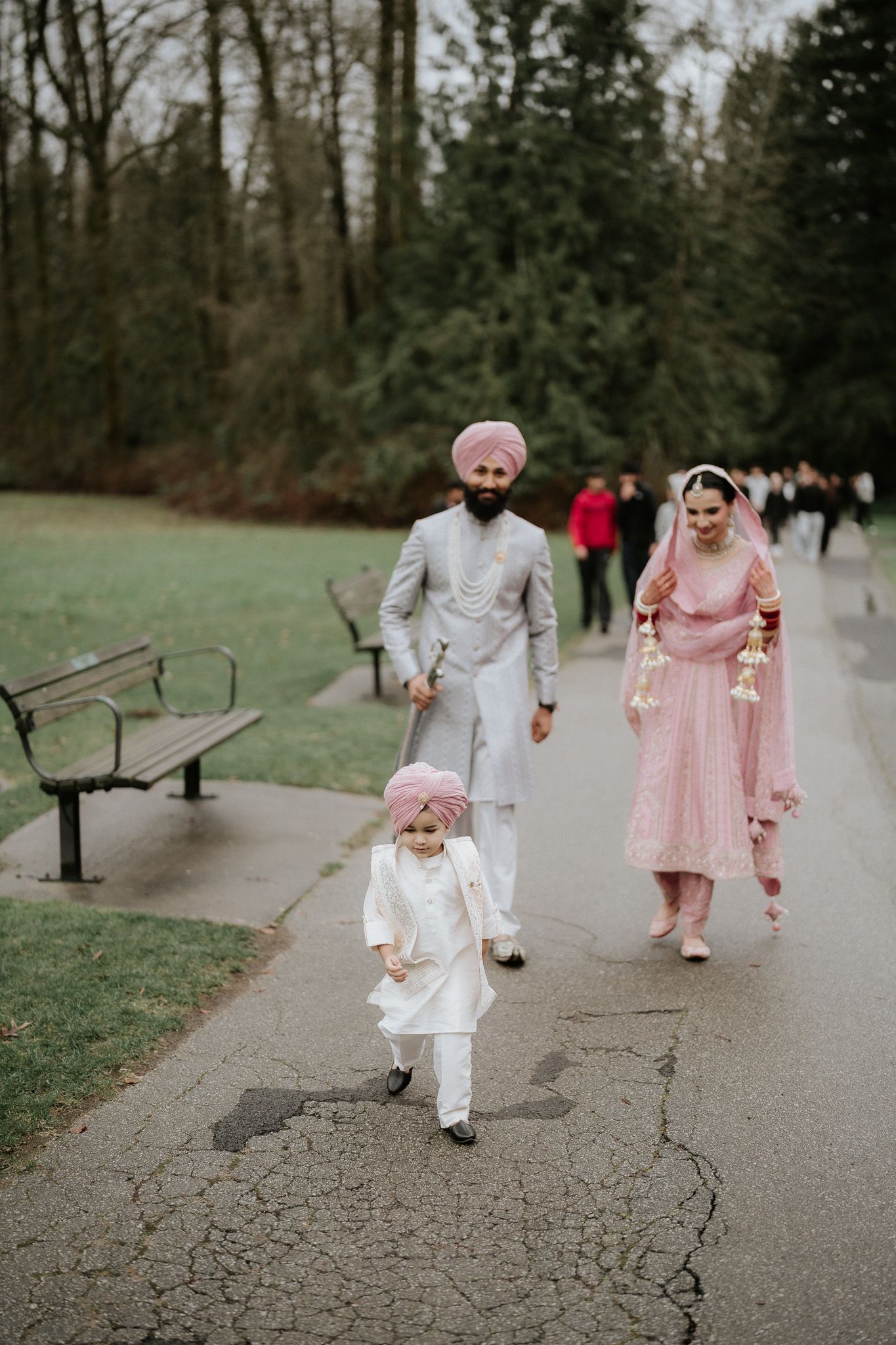 A family in traditional Indian wedding attire walks together on a park path with benches and trees in the background.