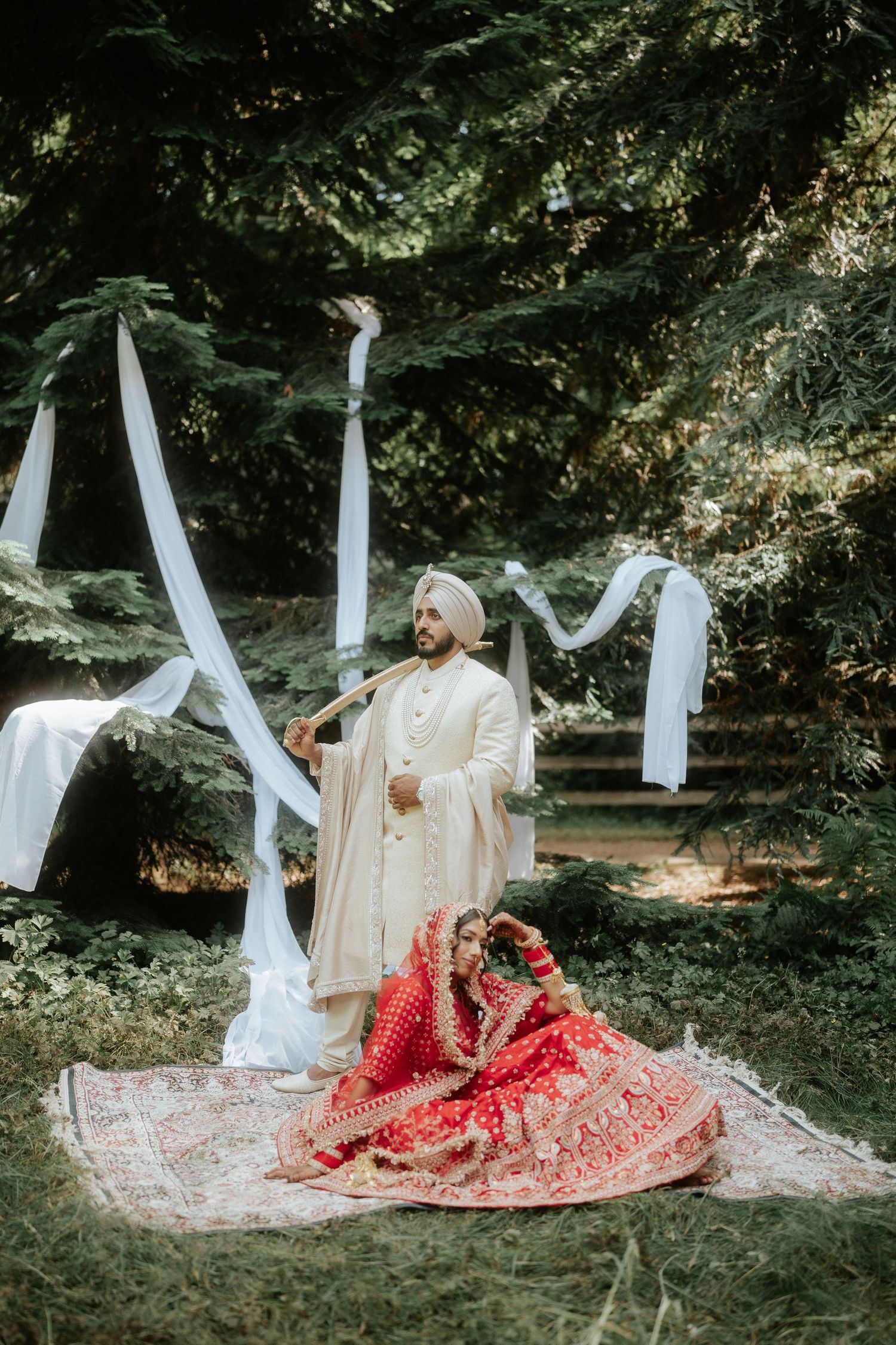 Wedding couple seated on decorative rug in forest with flowing white fabric installations.